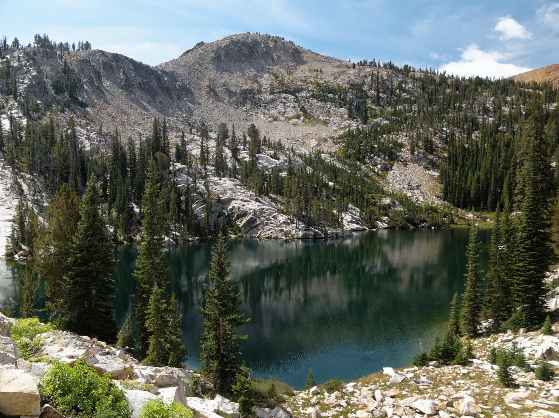Calm mountain lake reflecting pine trees and rocky slopes under a bright sky.
