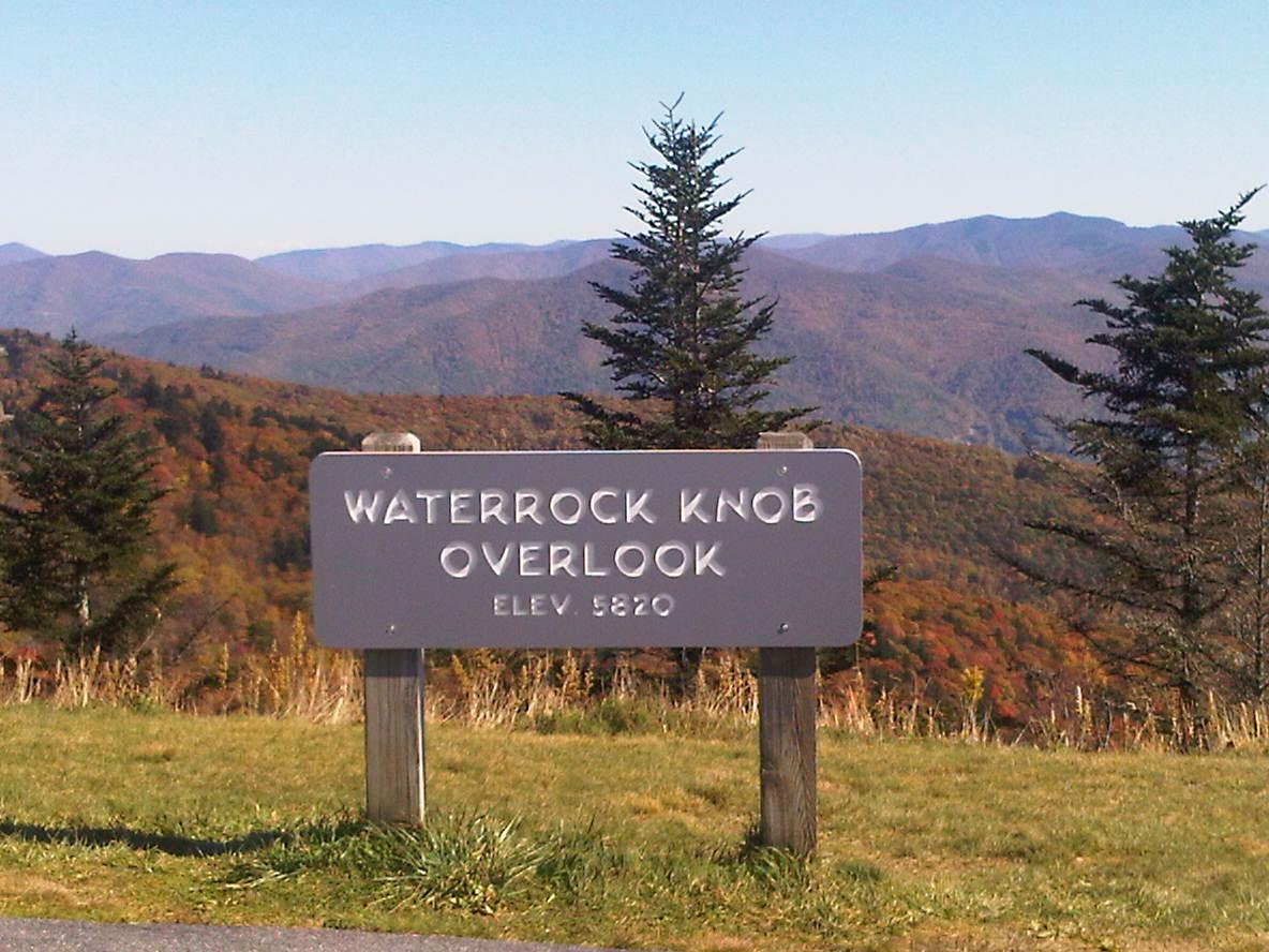 A wooden sign and evergreens against mountains displaying autumn colors under a clear sky.