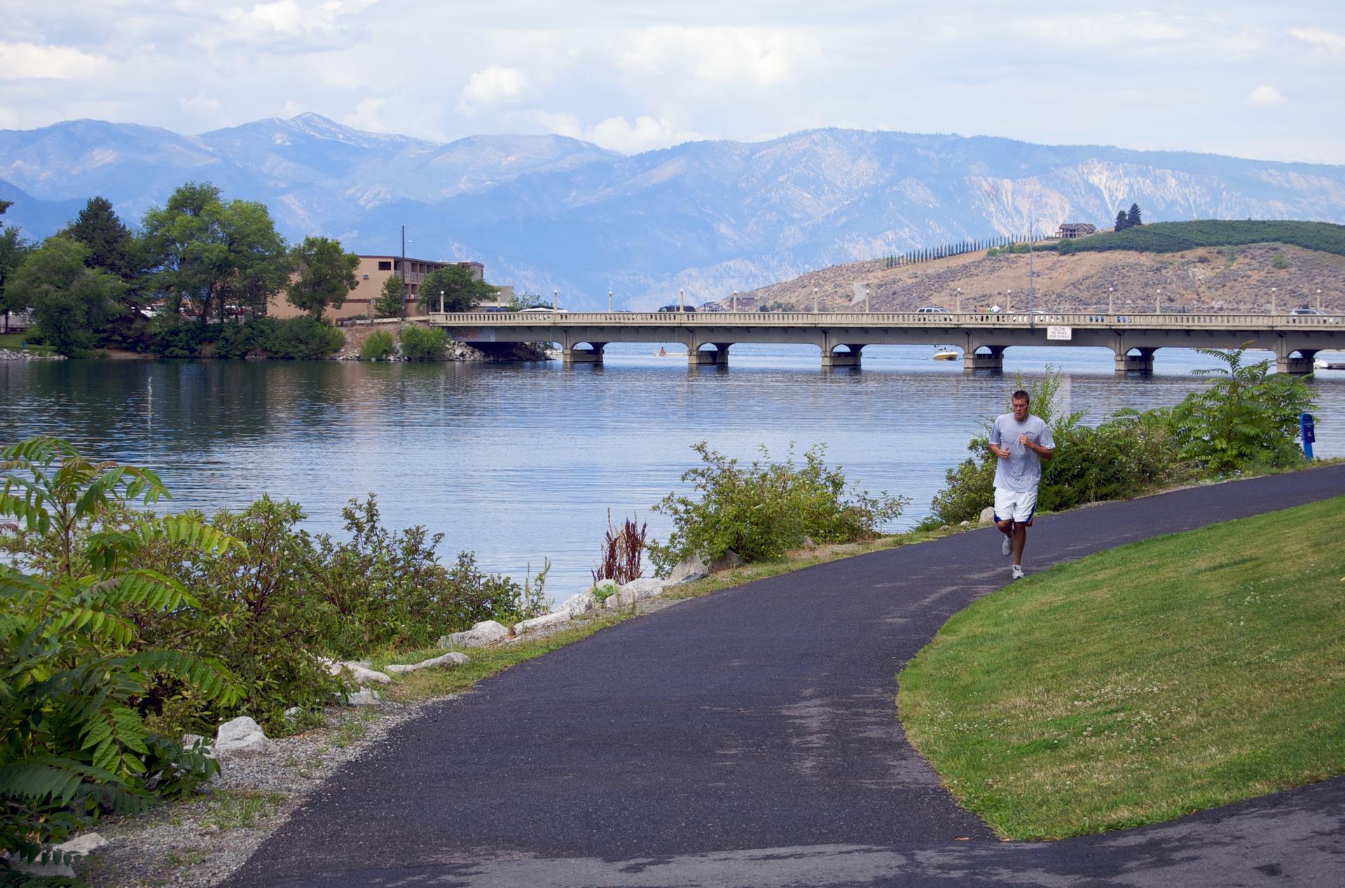 Man jogs on a paved path beside a lake with a bridge and mountains.