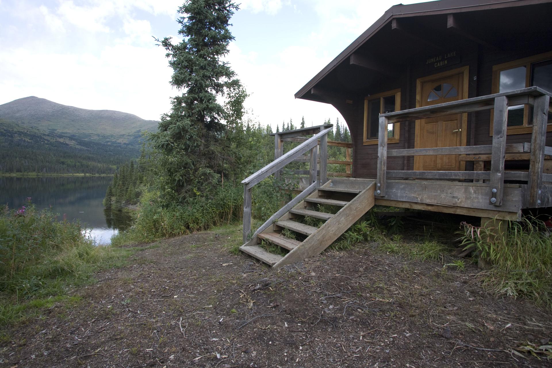 Wooden cabin with stairs next to a lake, mountains, and trees.