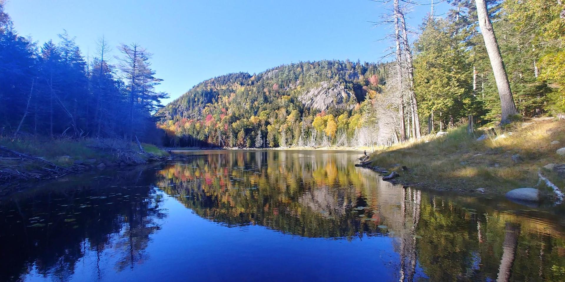 Serene lake reflecting autumn trees and a rocky hill on a clear day.