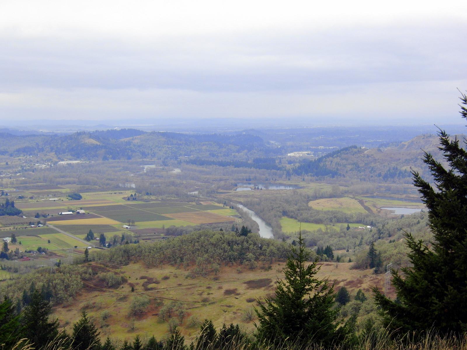 Expansive valley view with river, agricultural fields, and distant hills under overcast sky.