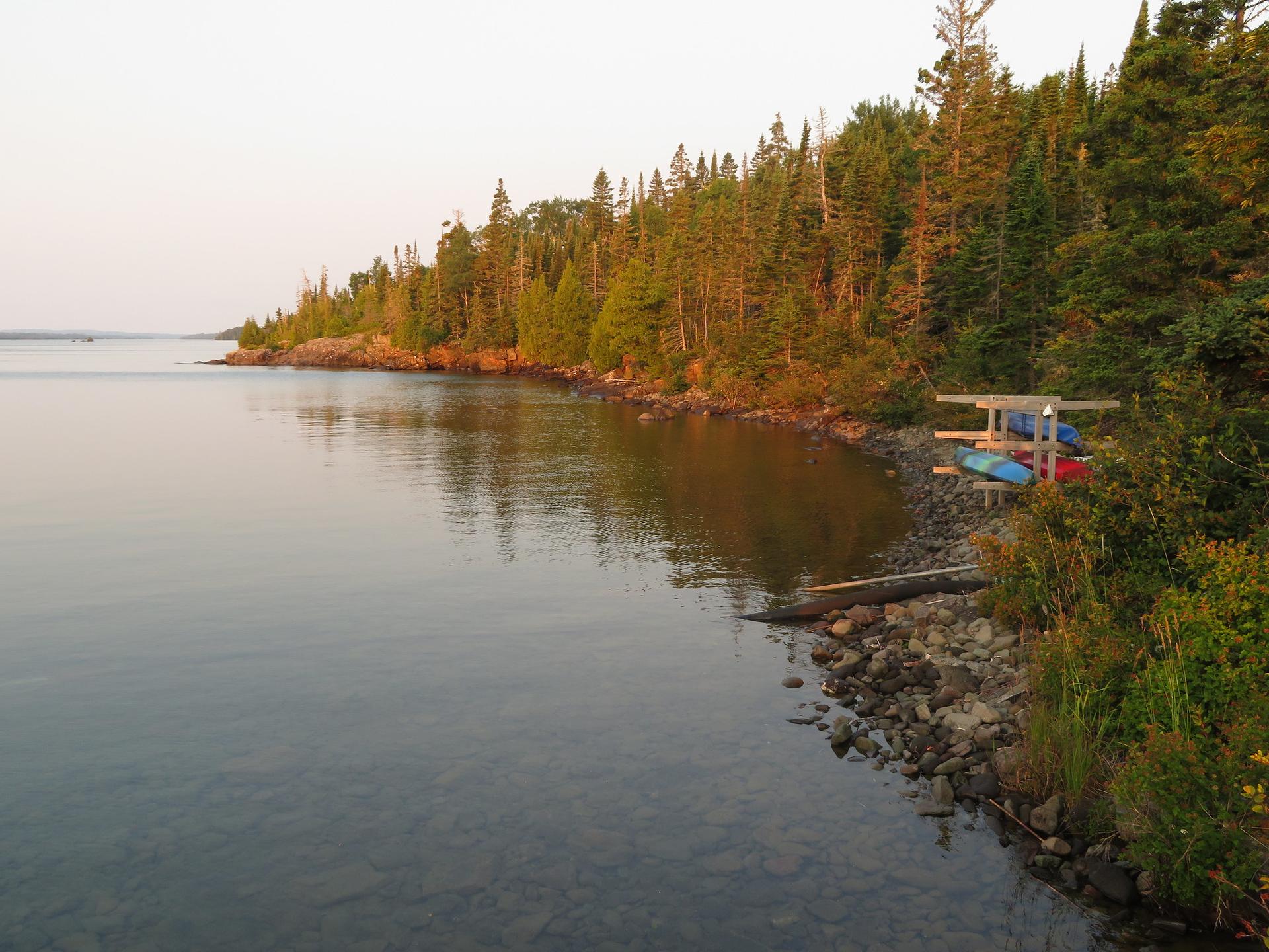Rocky shoreline with evergreen trees and canoes on a rack at sunset.