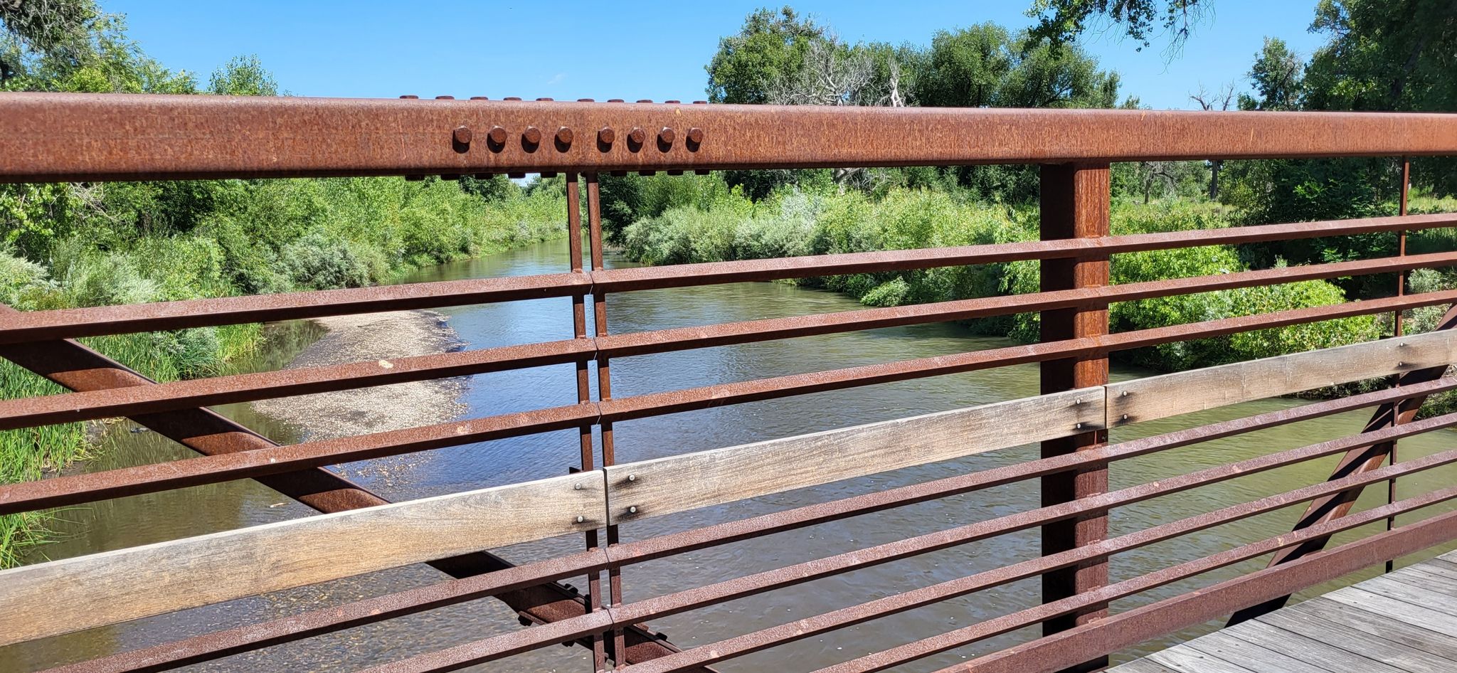Rusty metal bridge railing overlooking a river with green foliage. - Big Thompson River Trail