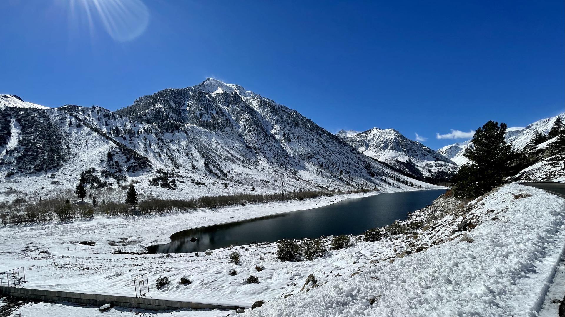 Snowy mountain landscape with a lake, bright sun, and clear blue sky.