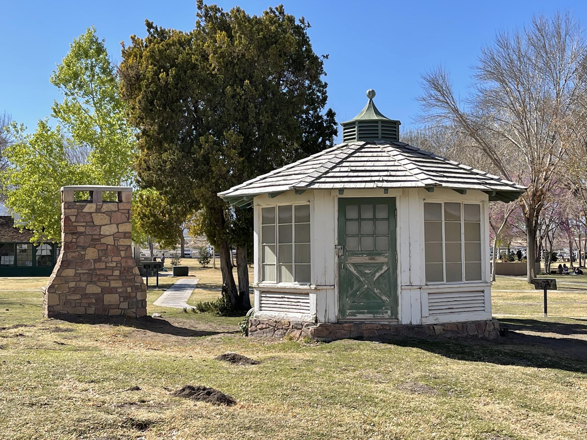 White gazebo with green door, stone chimney, and trees under a bright sky.