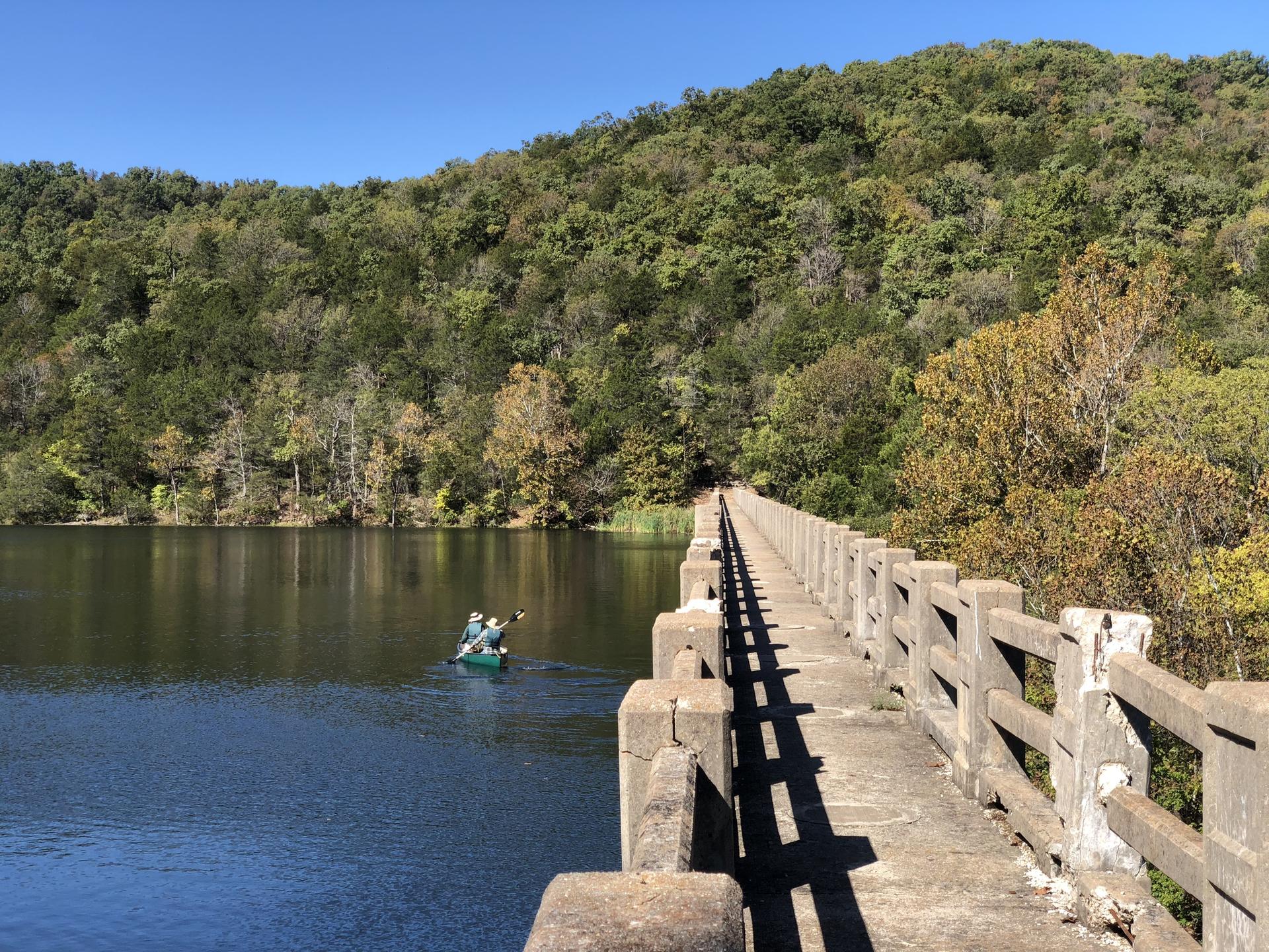 One person paddling a canoe near a long concrete bridge on a sunny day.