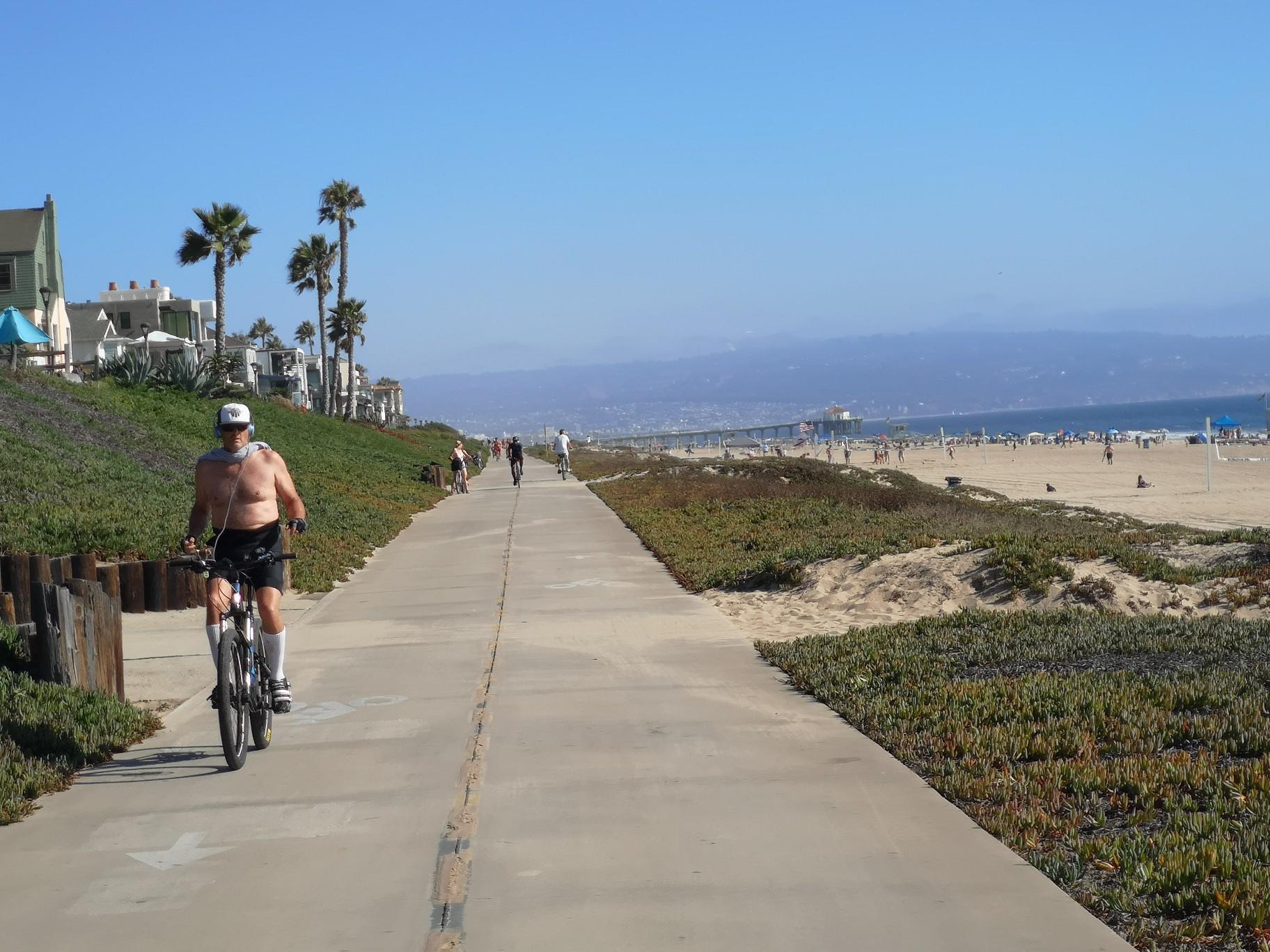 A man cycling on a paved beach path with people on the sandy beach, sunny.