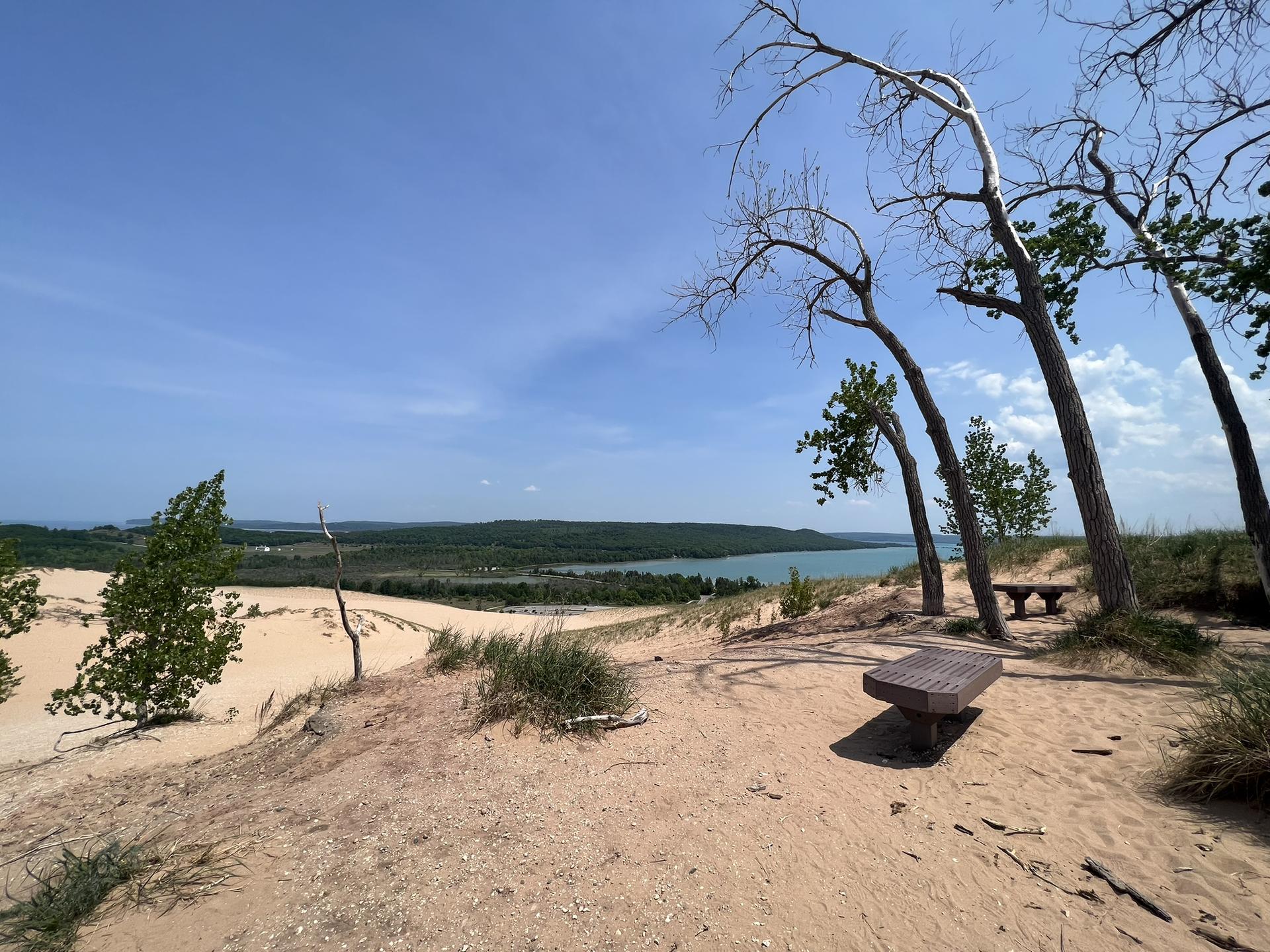 A bench on a sandy hill overlooking distant water and green landscape.