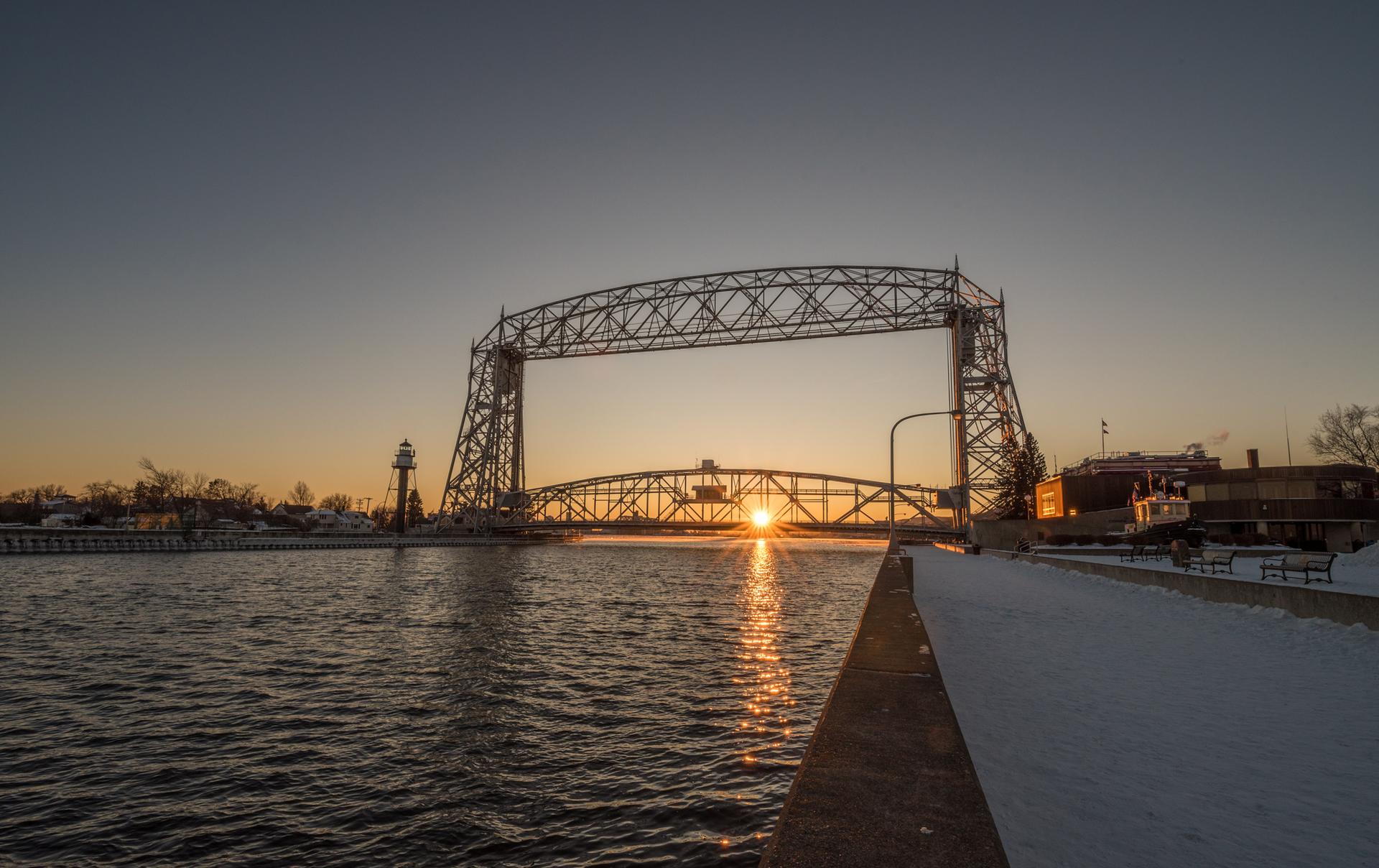 Sunset over a large metal bridge spanning water, with snow on the ground.