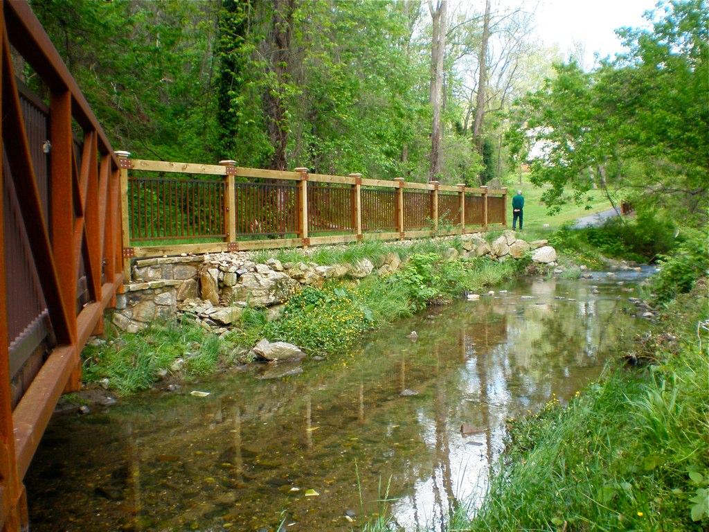 A wooden bridge and fence line a clear stream with a person in the distance.