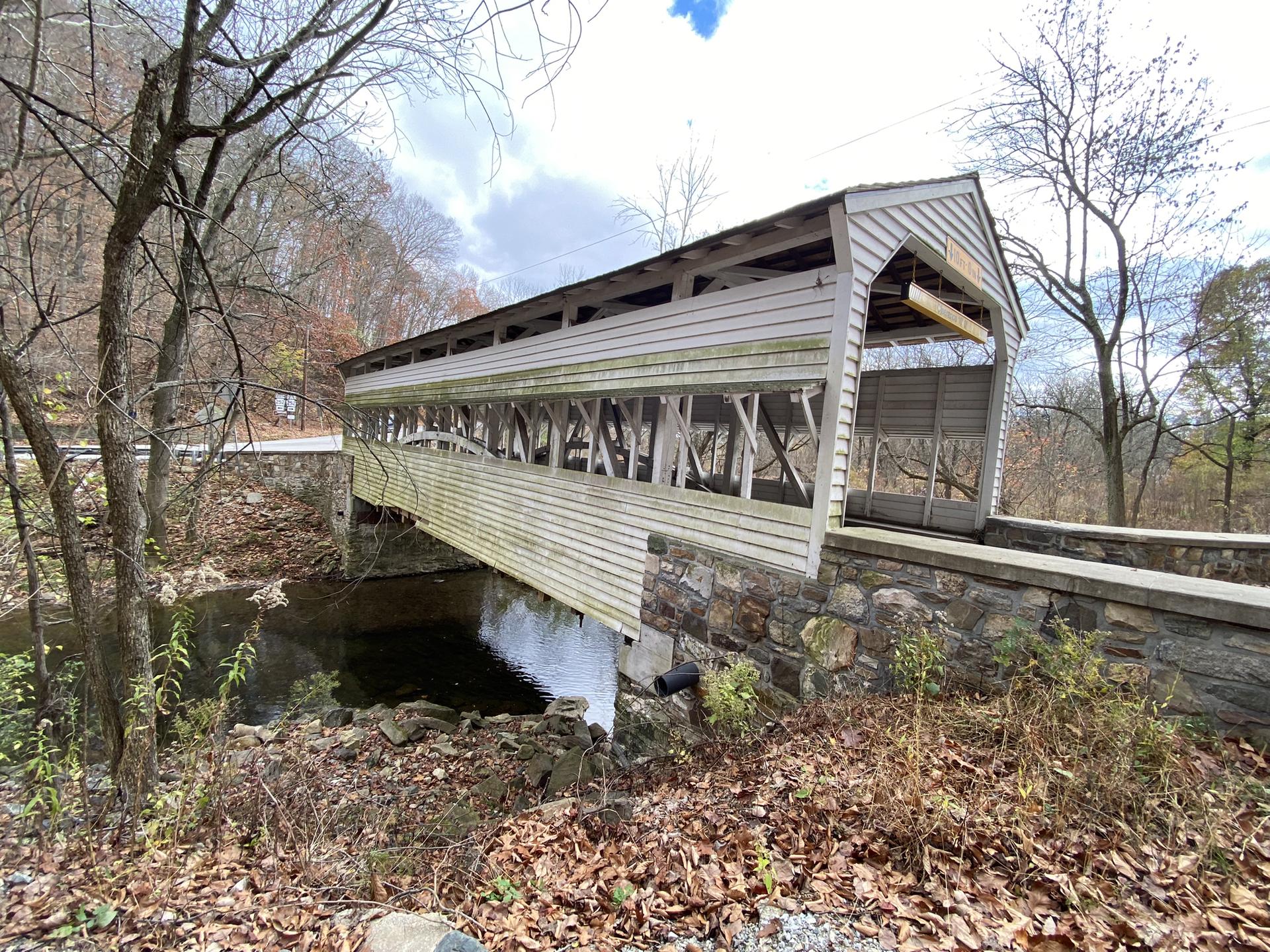 White covered bridge over a dark stream, surrounded by autumn trees.