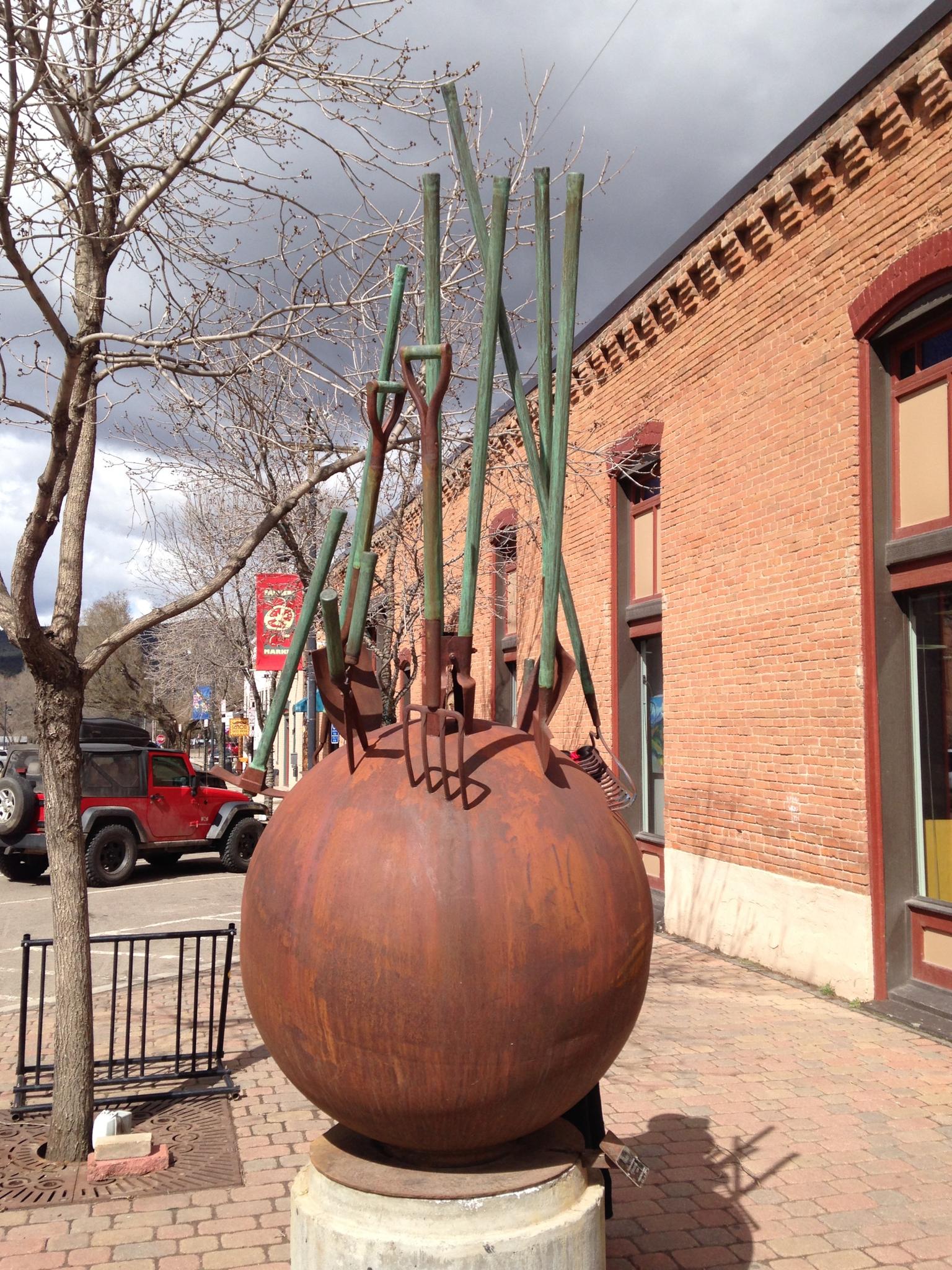 Large rusty sphere sculpture with garden tools on a sunny urban sidewalk.