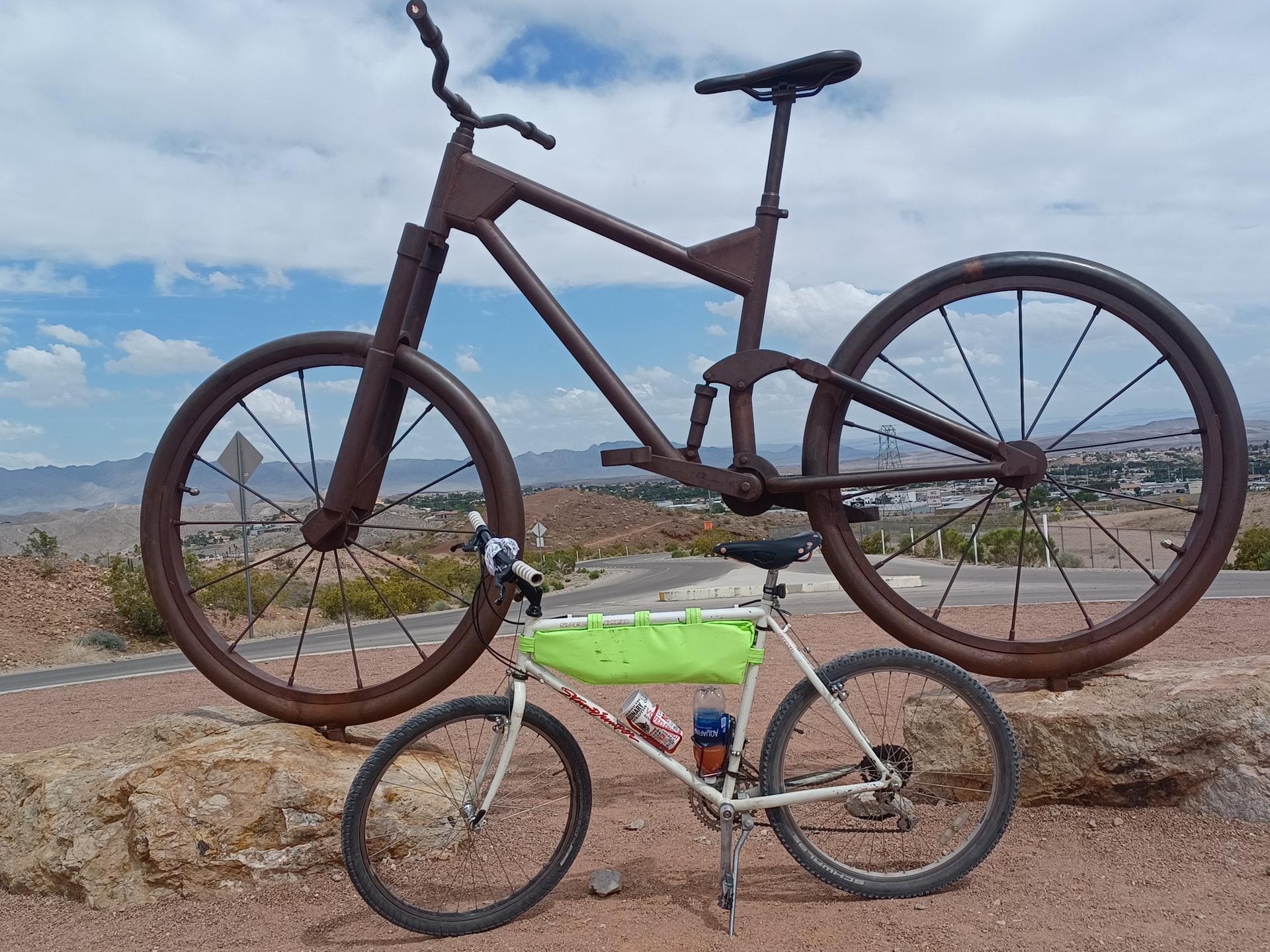 A small bicycle rests before a giant metal bicycle sculpture outdoors.
