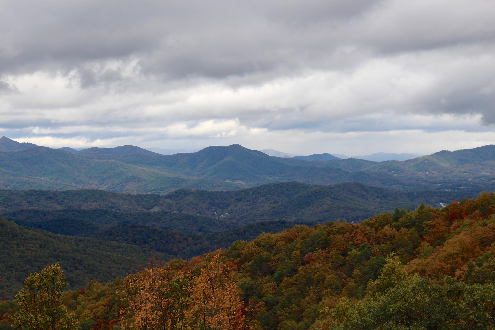 Overcast sky above rolling mountains with autumn-colored trees in foreground.