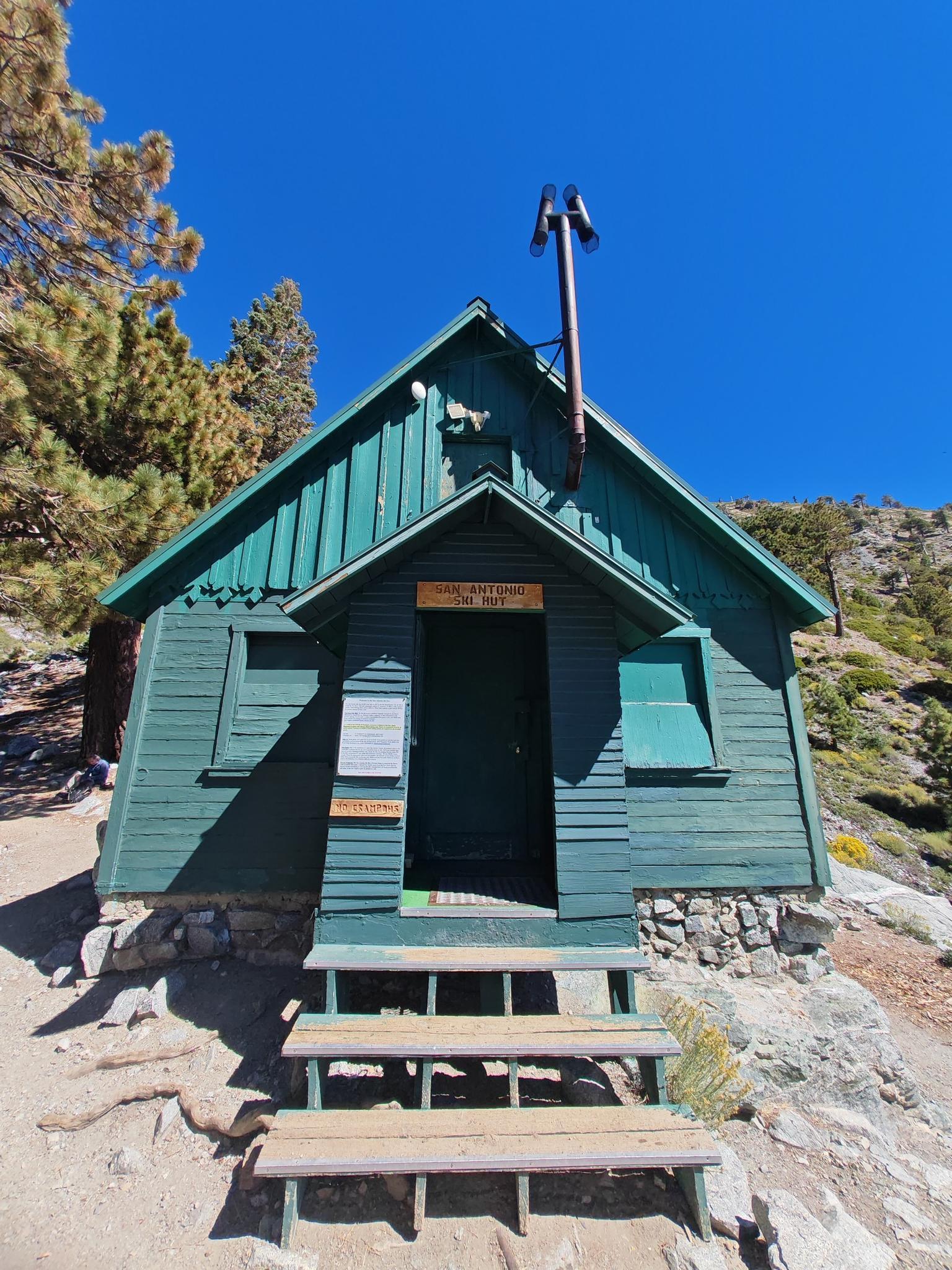 A green wooden cabin with a porch and chimney under a clear blue sky.