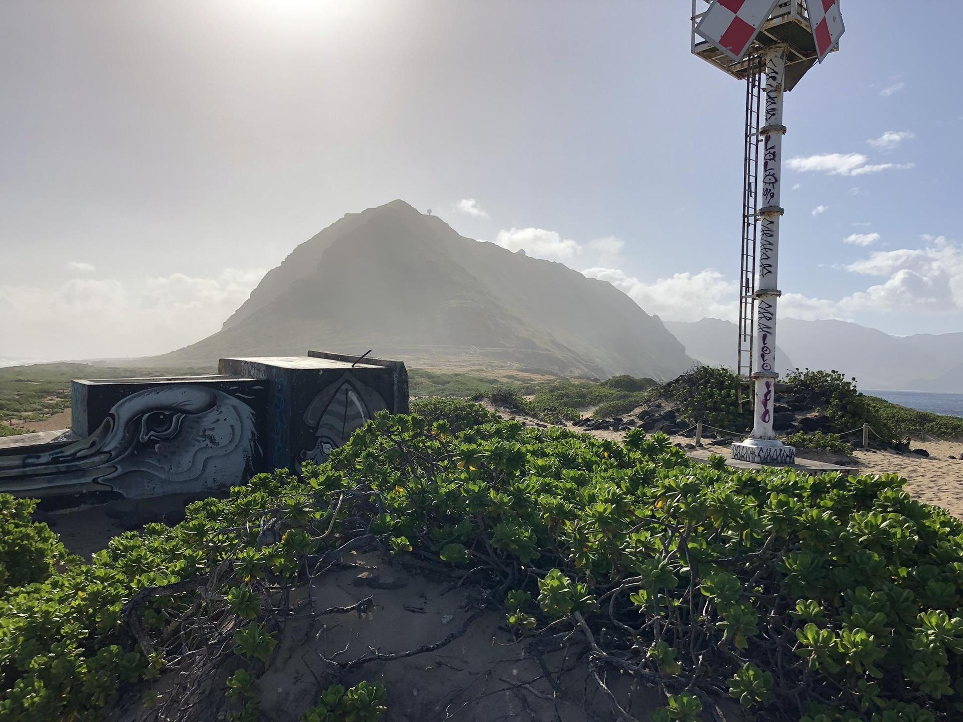 Bright sun over a mountain, with a red and white tower and green bushes.