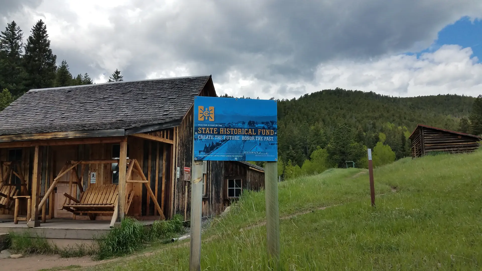 Rustic wooden buildings and a blue sign on a grassy hill under a cloudy sky.