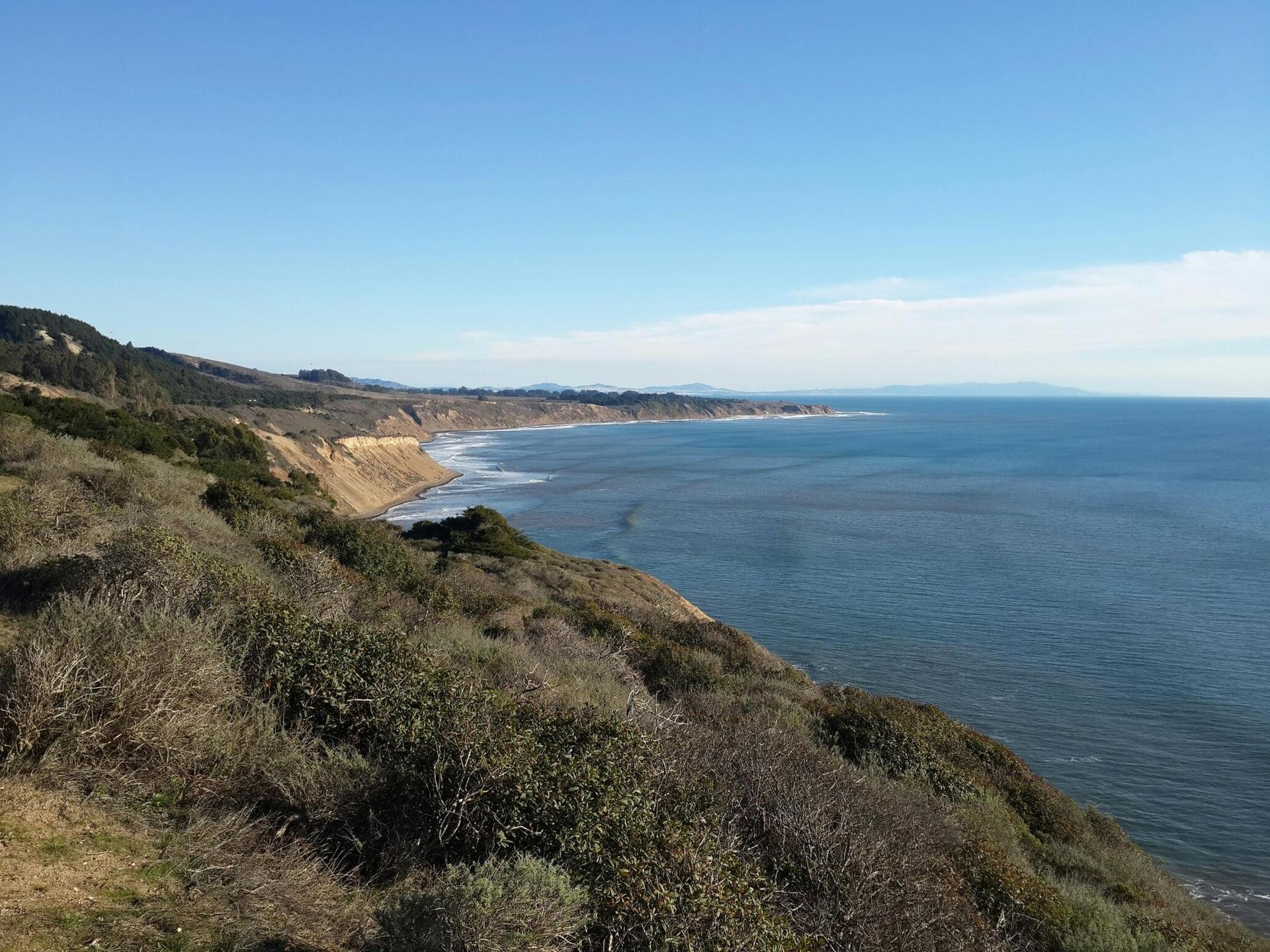 Sunny coastal cliffs overlooking a vast blue ocean with distant land.