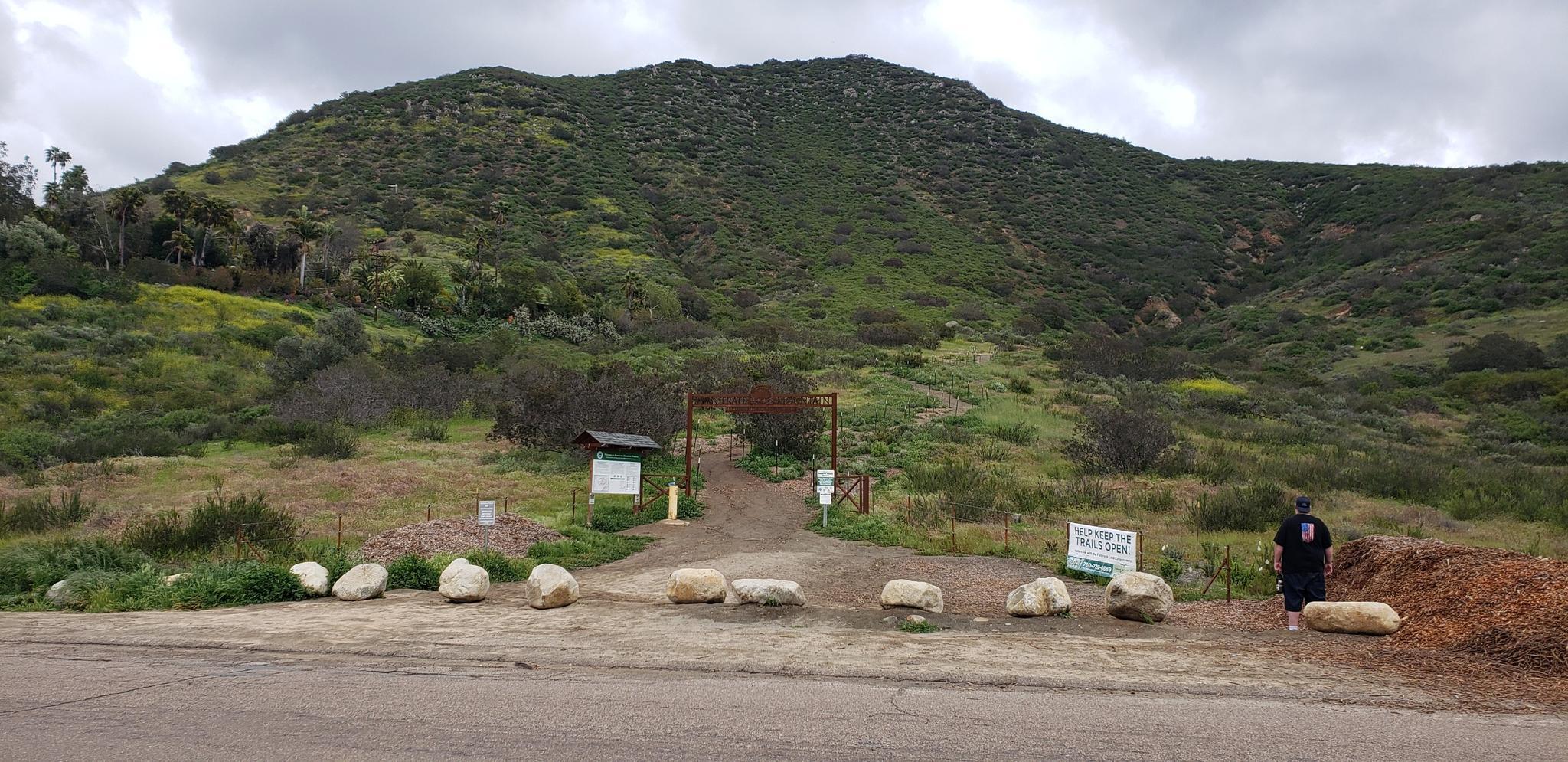 A person stands at a trailhead entrance with a large green hill.