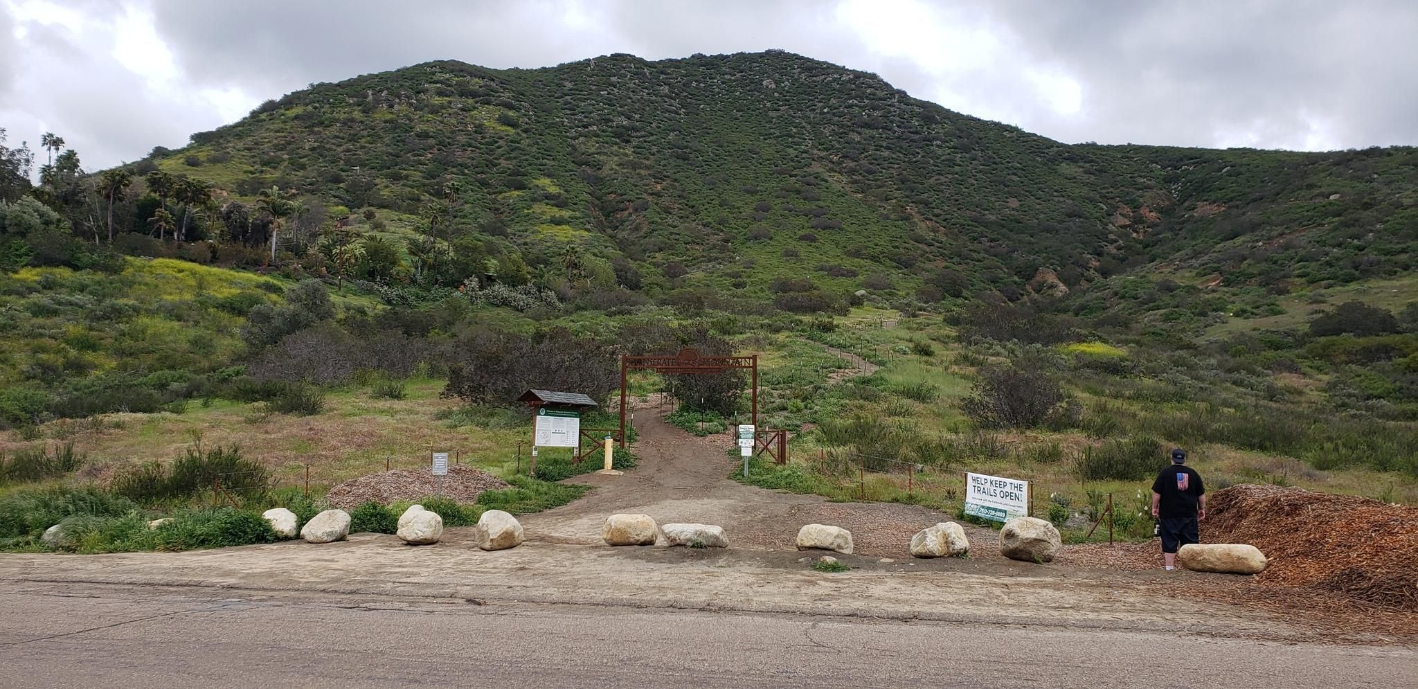 A person stands at a trailhead entrance with a large green hill. - Monserate Mountain Loop Trail