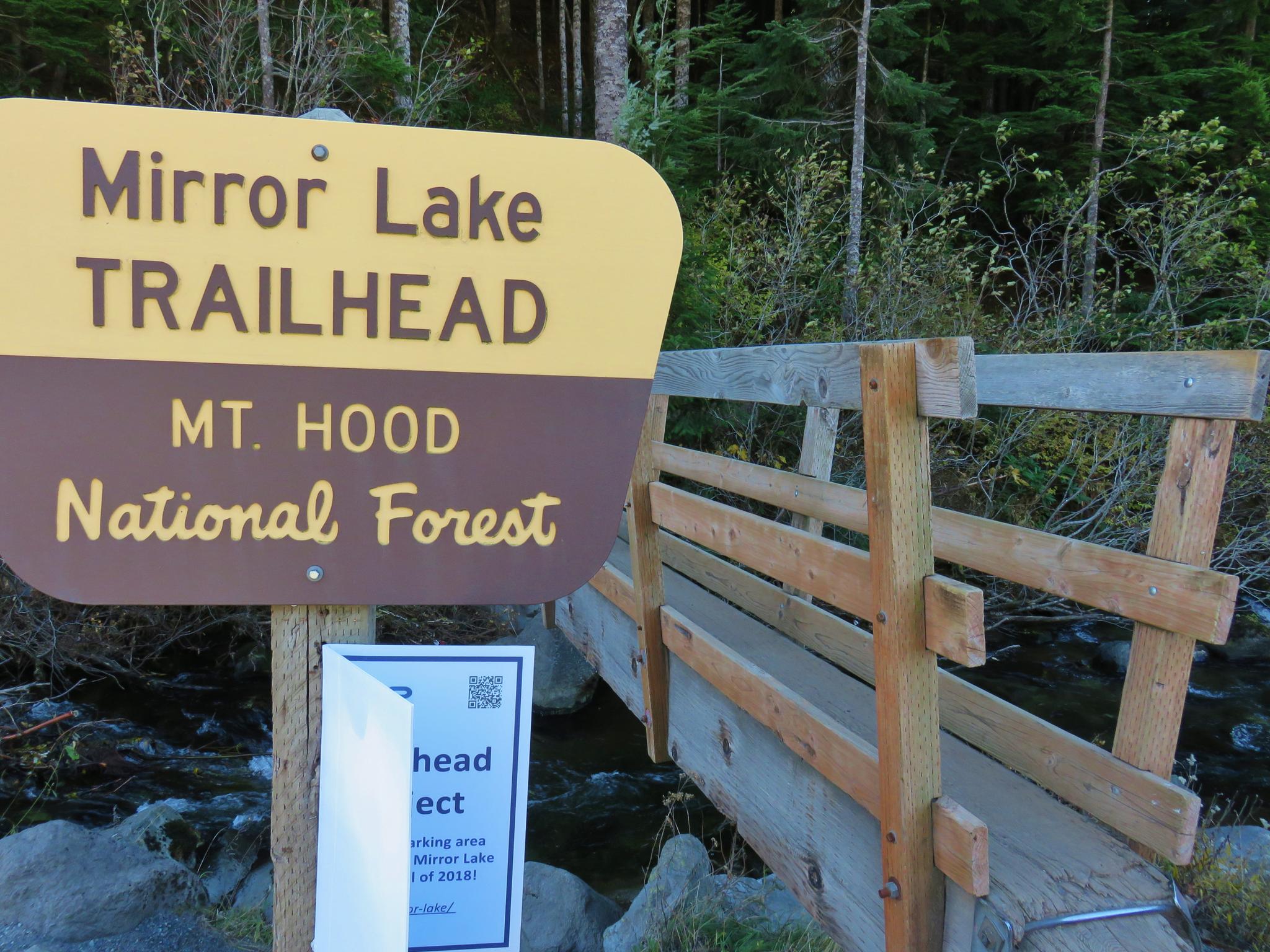 A trailhead sign next to a wooden bridge over a stream in a forest.