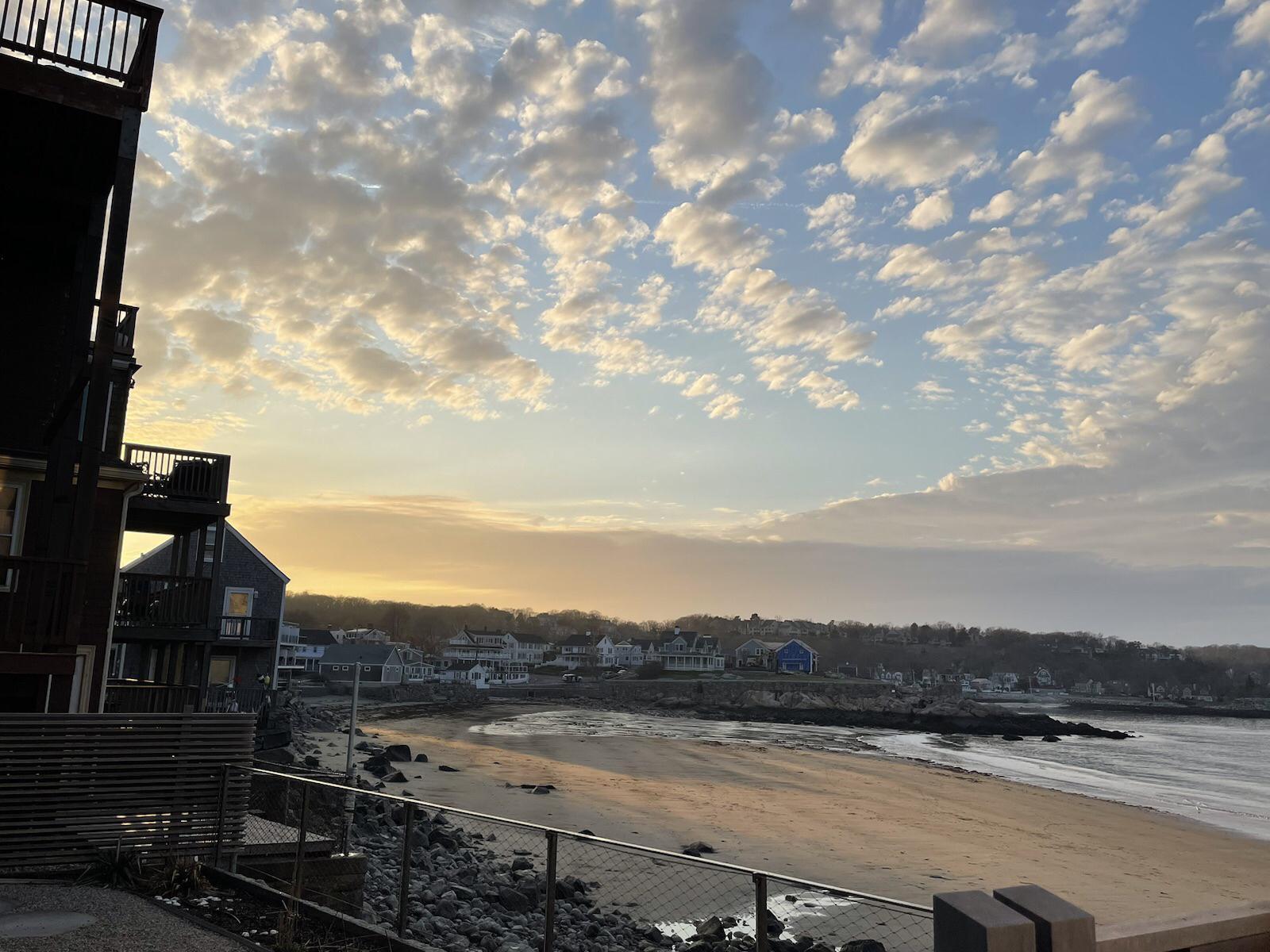 Golden hour light illuminates a sandy beach and coastal buildings under a cloudy sky.