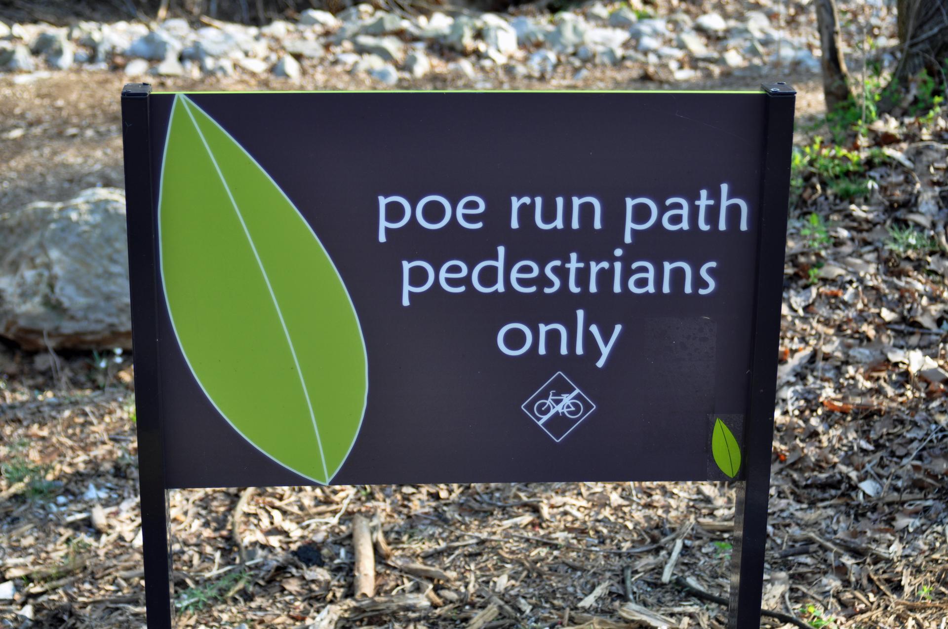 A black sign with a green leaf, stating "pedestrians only" on a path.