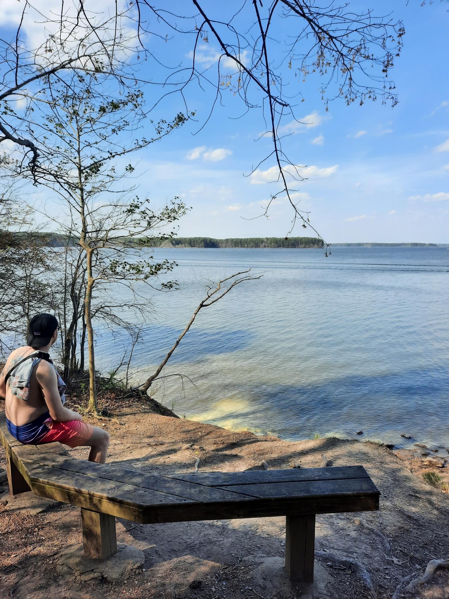 A person sits on a wooden bench overlooking a vast lake.