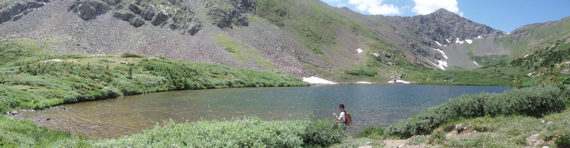 Person by clear mountain lake surrounded by green hills under blue sky.