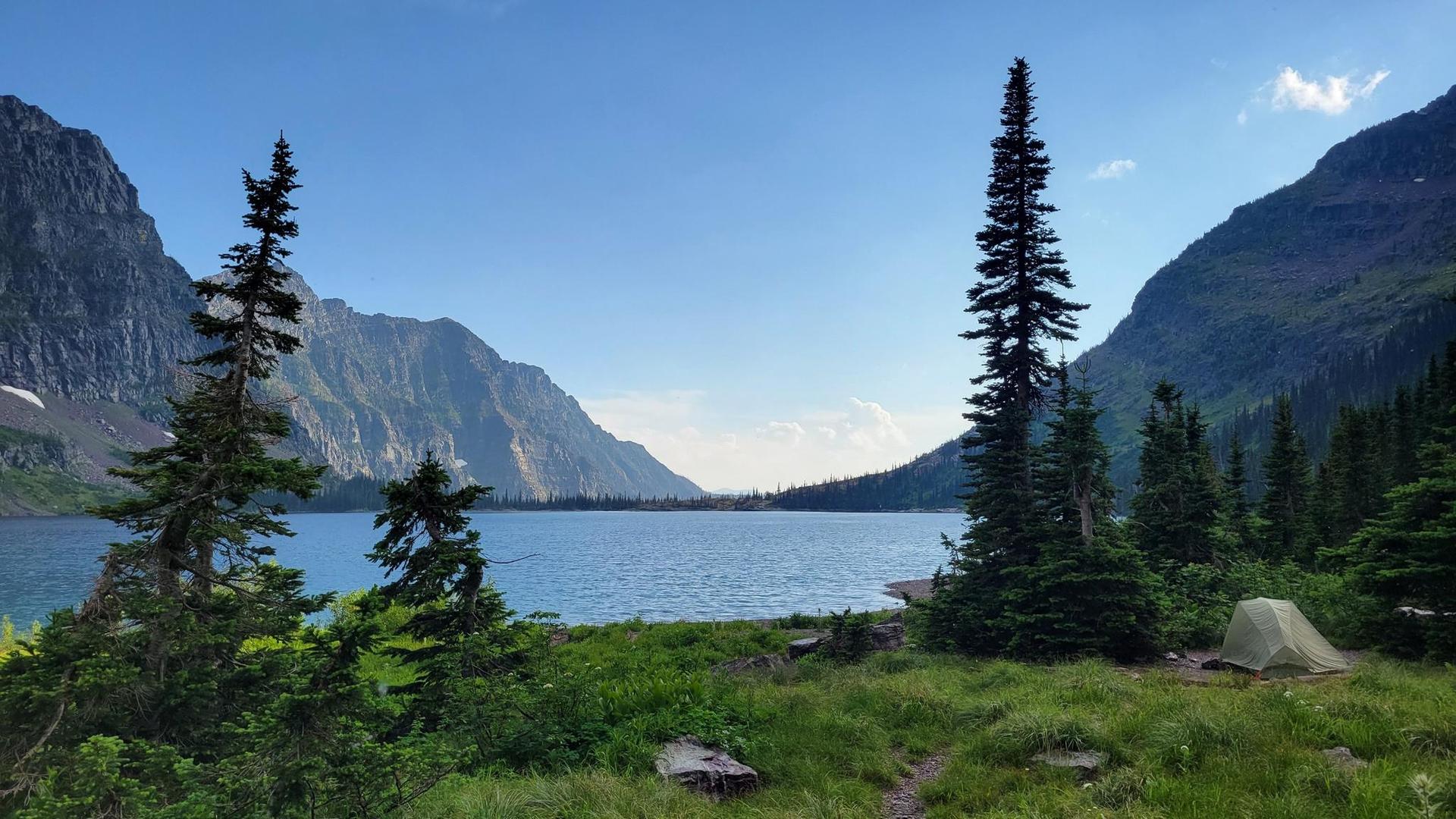 A tent sits by a large mountain lake surrounded by towering peaks and trees.