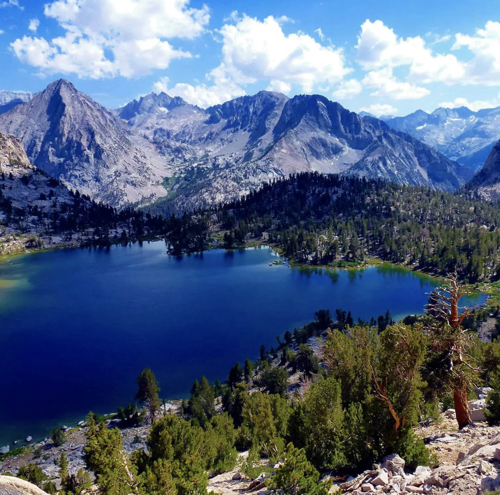 Deep blue mountain lake surrounded by rocky peaks and pine forests under a bright sky. - Monarch Lakes Trail