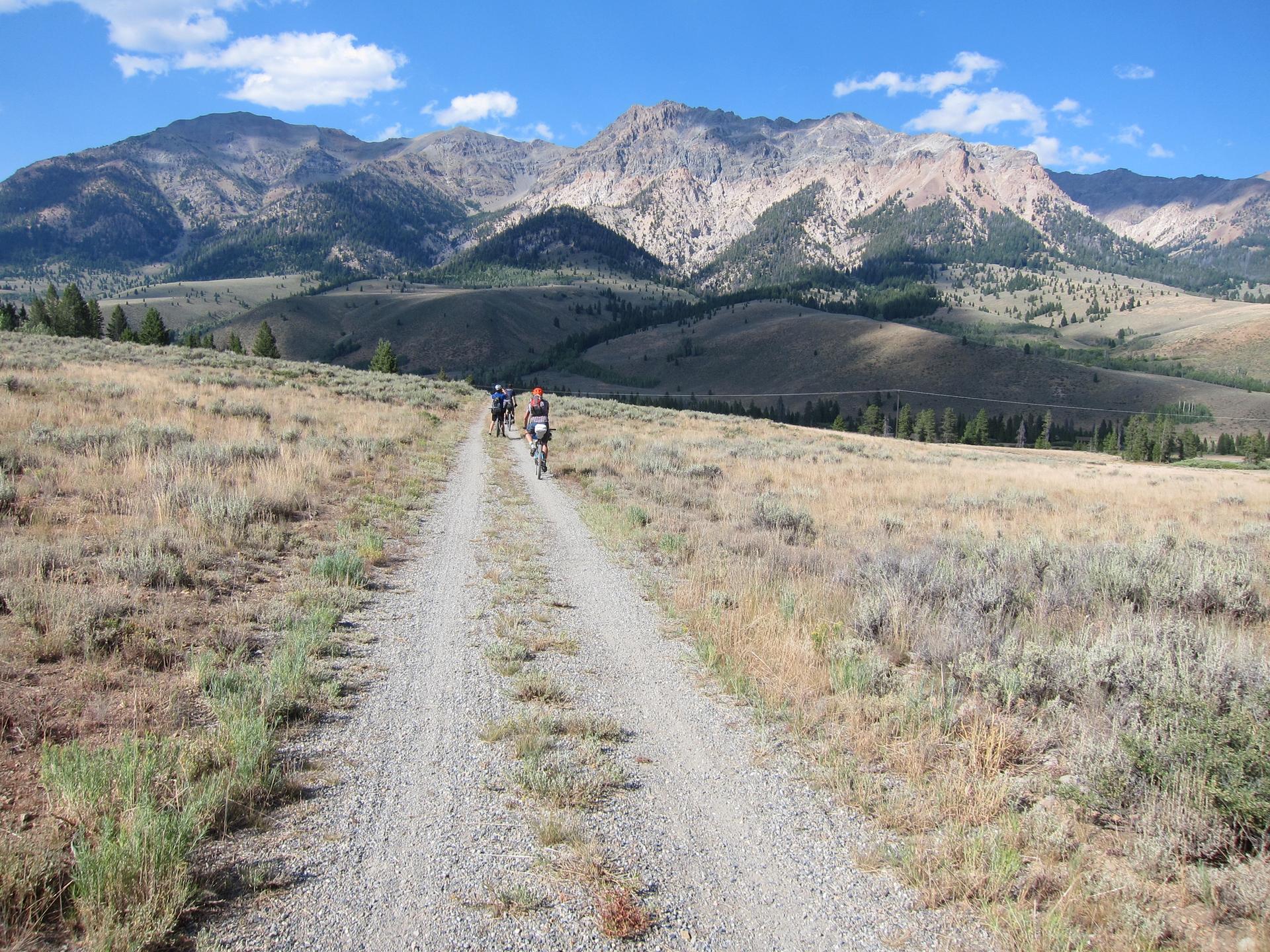 Two cyclists ride a gravel road through a dry, hilly landscape with distant mountains.