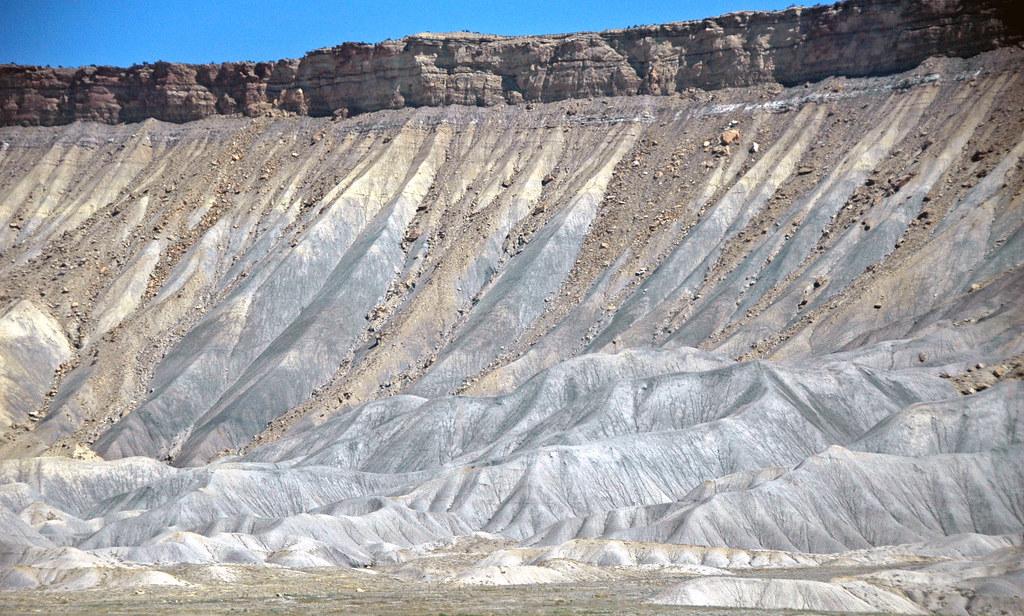 Dramatic layered rock formation with steep, eroded slopes under a bright blue sky.