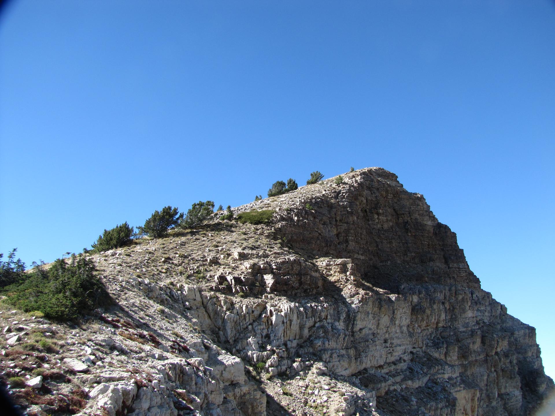 Steep rocky mountain peak with sparse vegetation under a bright blue sky.