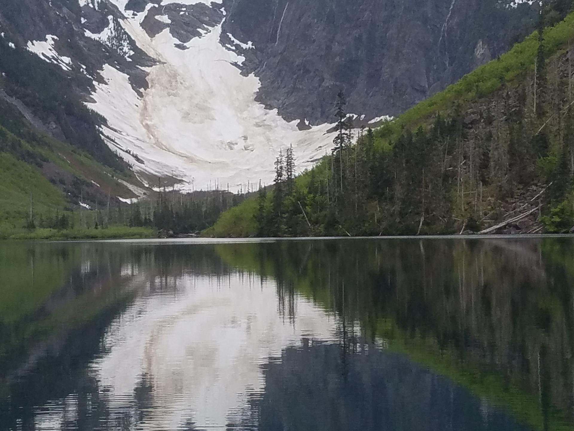 Calm mountain lake reflecting snow-covered peaks and green forests.
