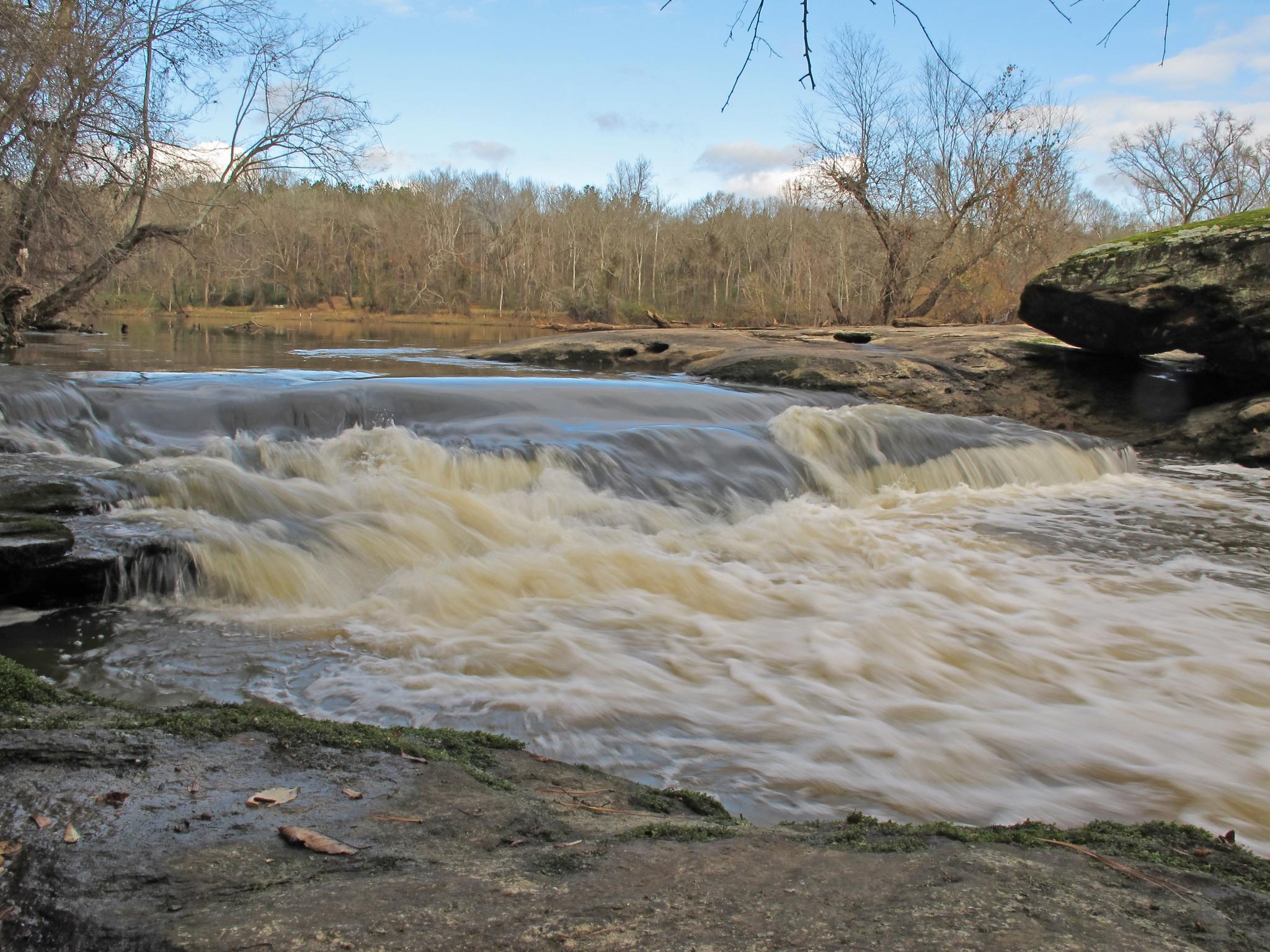 Rushing river rapids flow over rocks under a cloudy sky.
