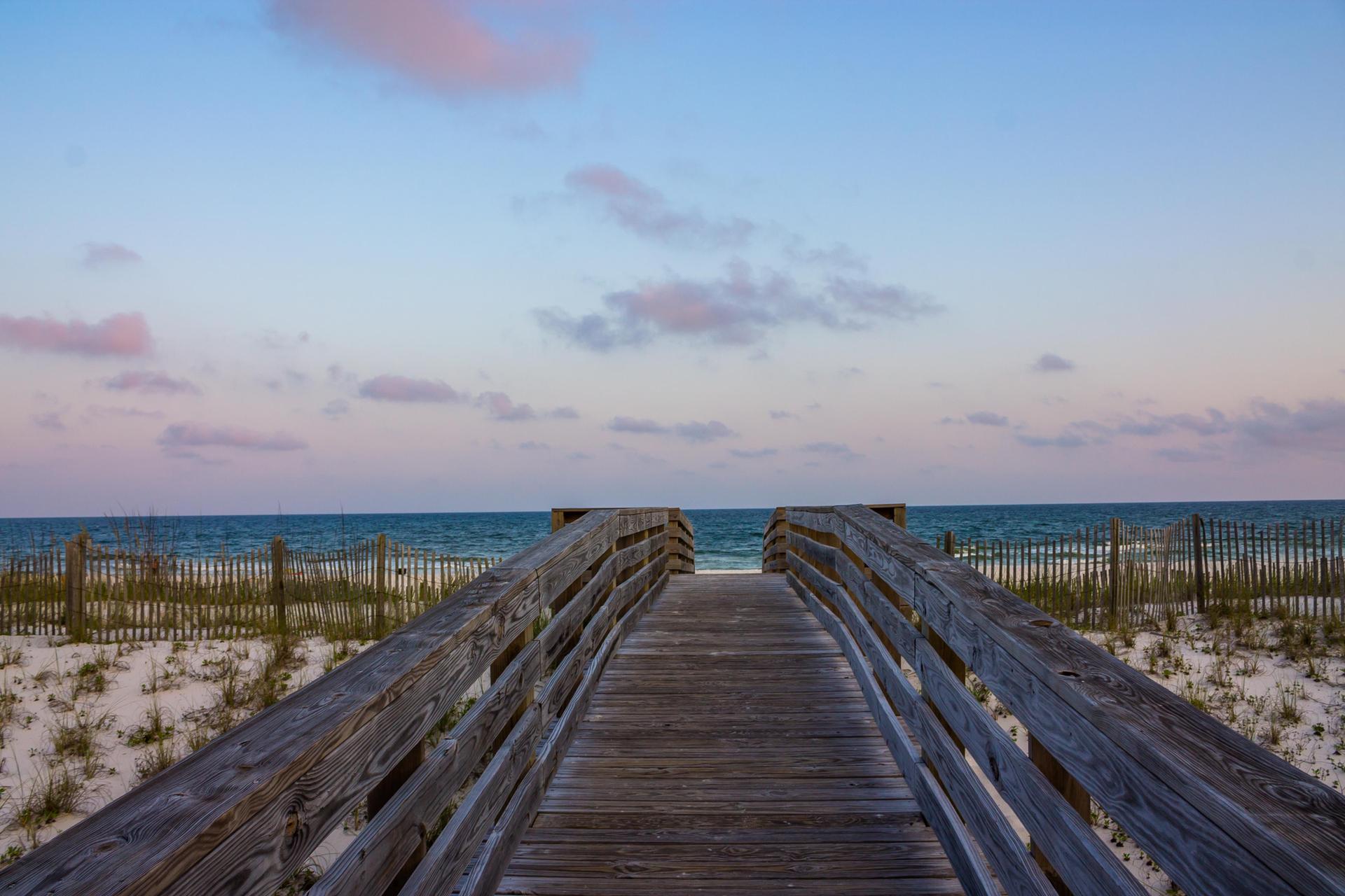 Wooden boardwalk leading to a sandy beach and ocean under a colorful sky.