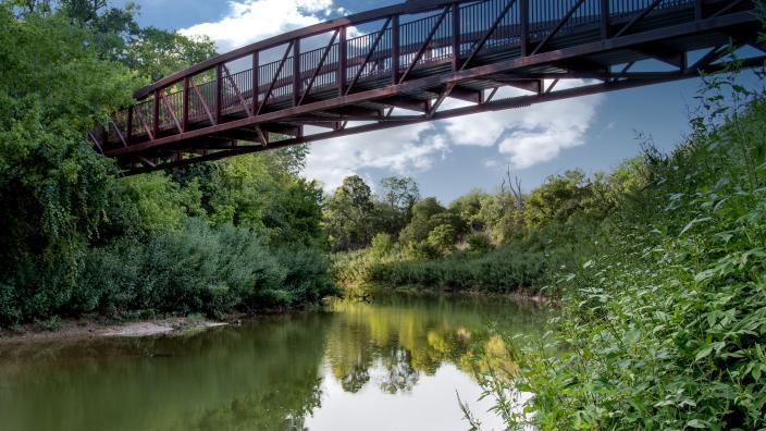 A sturdy metal bridge crosses a calm river surrounded by dense foliage.