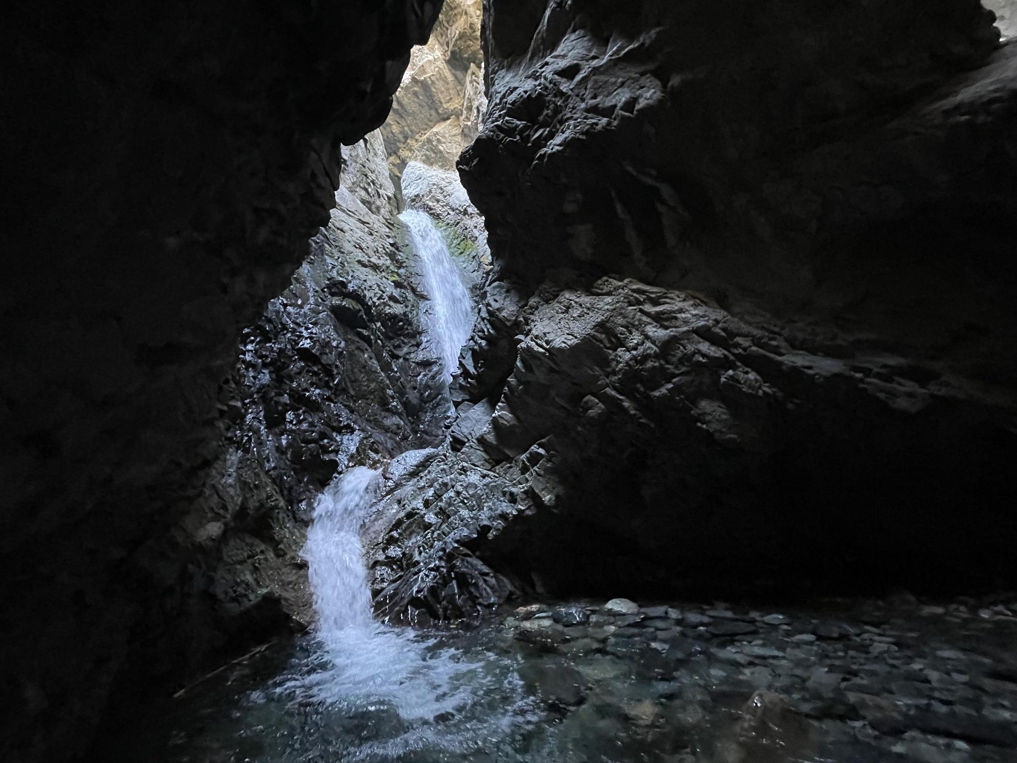Waterfall cascades into a dark, narrow, rocky canyon with flowing water.