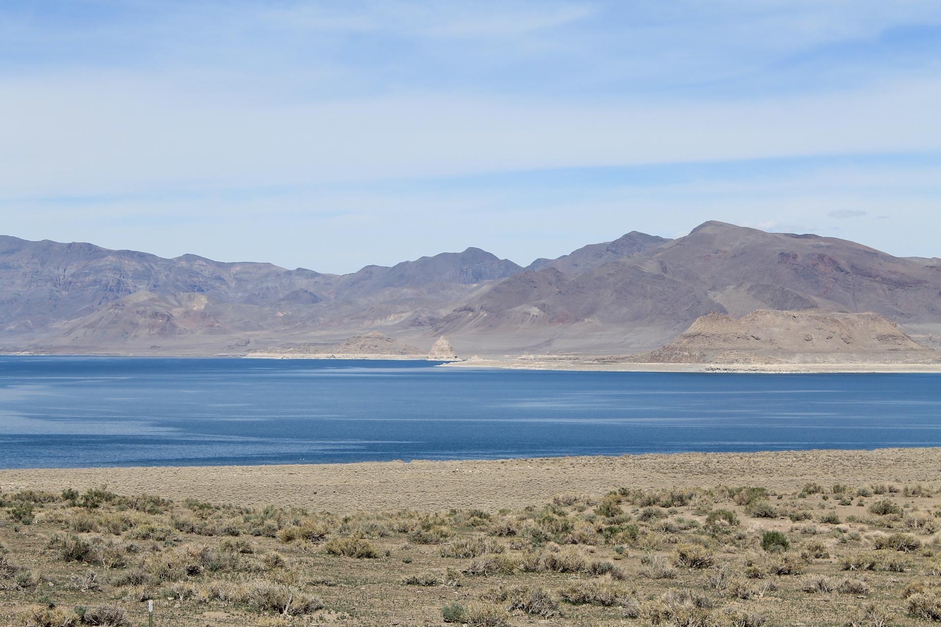 Blue lake surrounded by dry, brushy land and distant, rocky mountains under a bright sky.