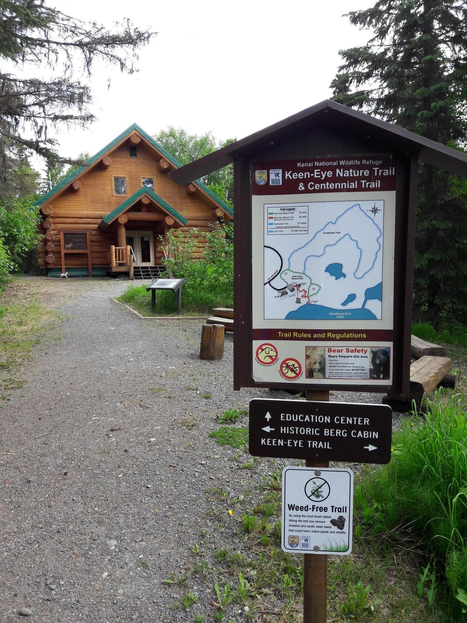 Trailhead sign and log cabin in a wooded area on an overcast day.