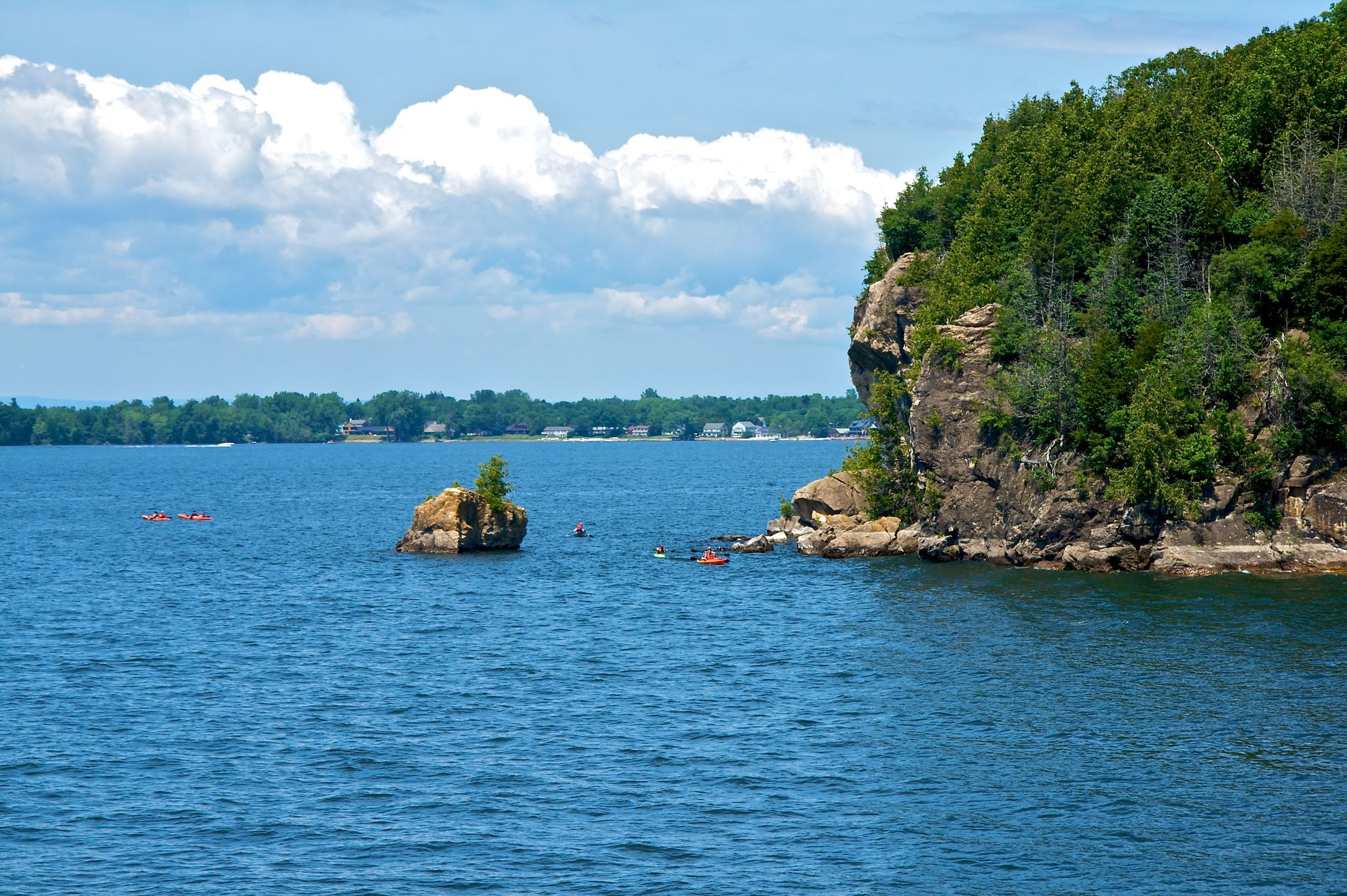 Kayakers paddle near a rocky island and forested cliff on a sunny day. - Rock Point Trail Loop