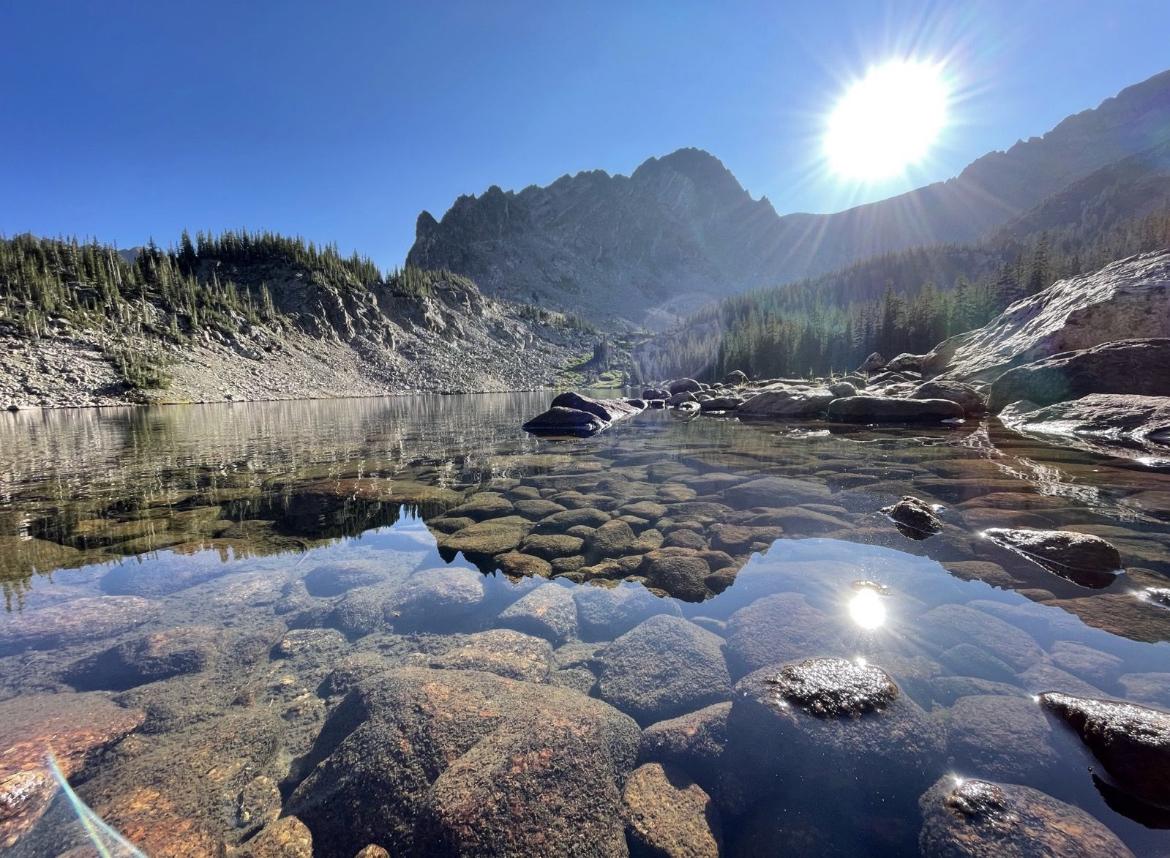 Clear mountain lake with visible rocks, surrounded by mountains under bright sun.
