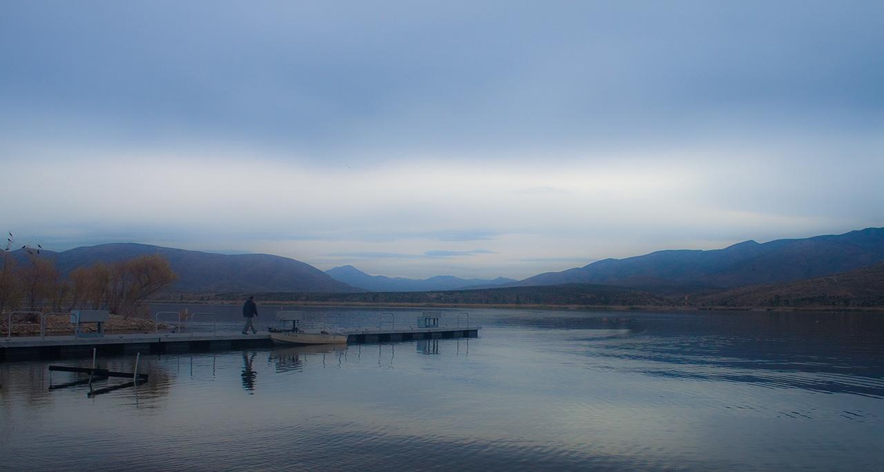 A person walks on a dock by a calm lake under an overcast sky.