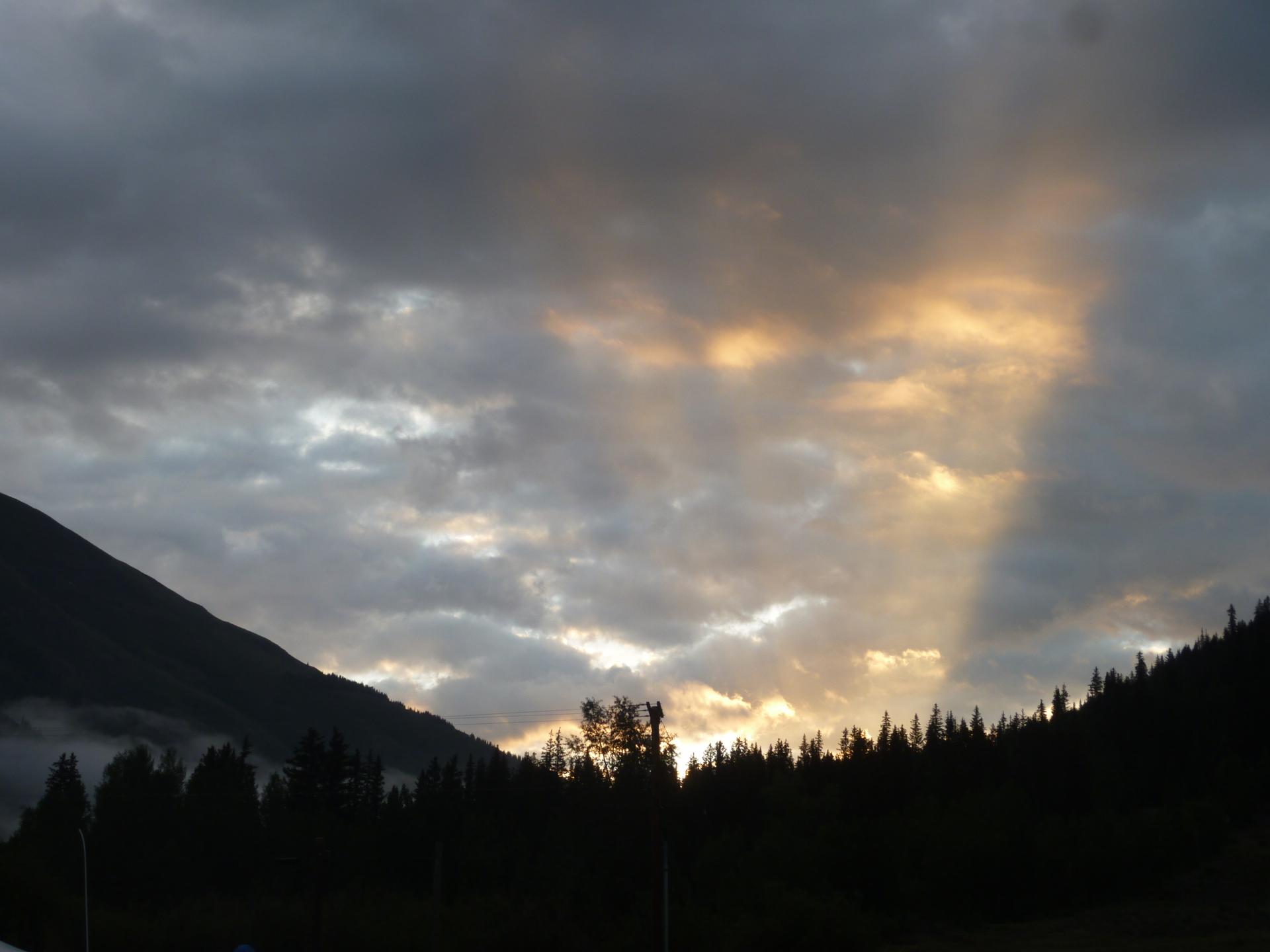 Dramatic sunset over dark forested mountains with sun rays breaking through clouds.