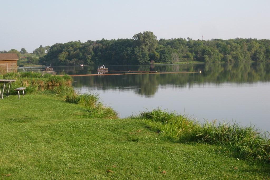 Calm lake with a grassy bank, distant trees, and people on a long pier.
