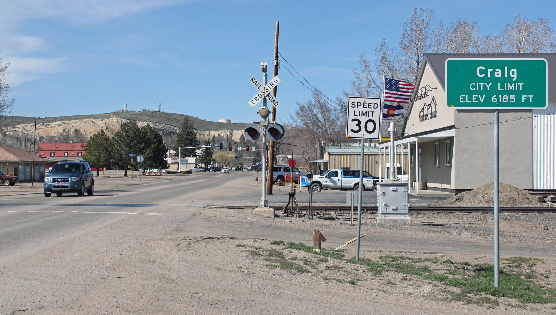 A sunny roadside with signs, a railroad crossing, buildings, and distant hills.