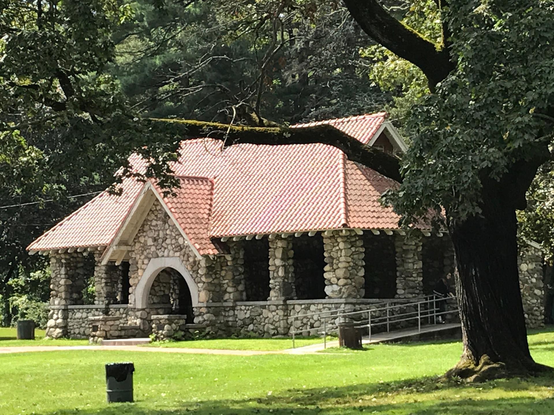 Stone pavilion with red roof surrounded by green grass and large trees in daylight.