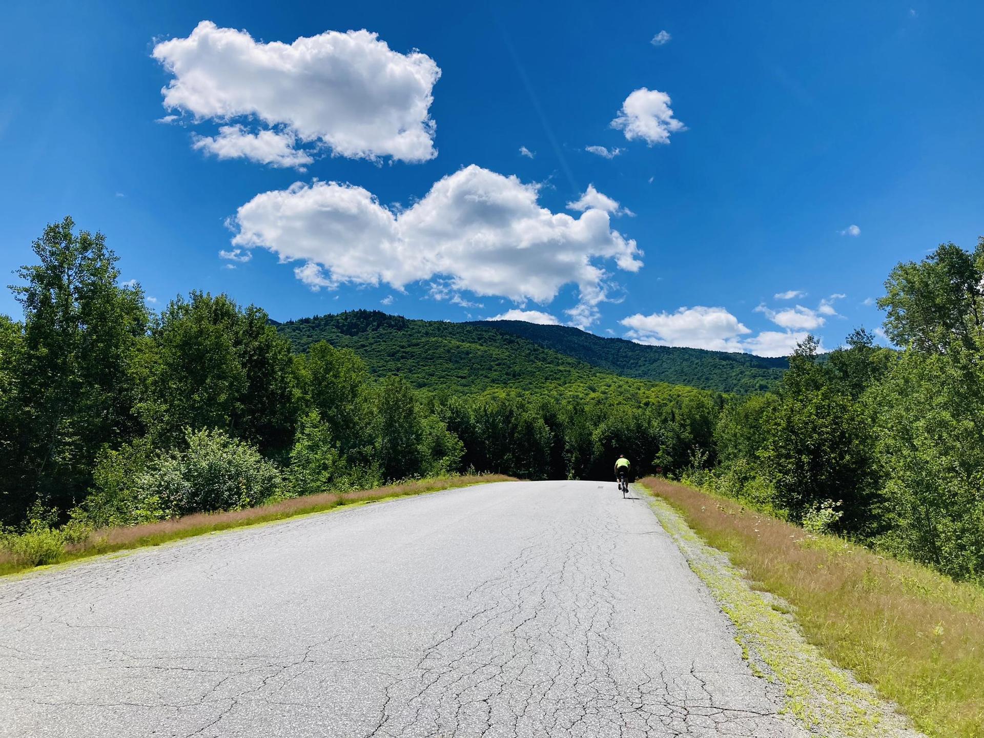 Cyclist on a paved road with lush green mountains under a bright blue sky.